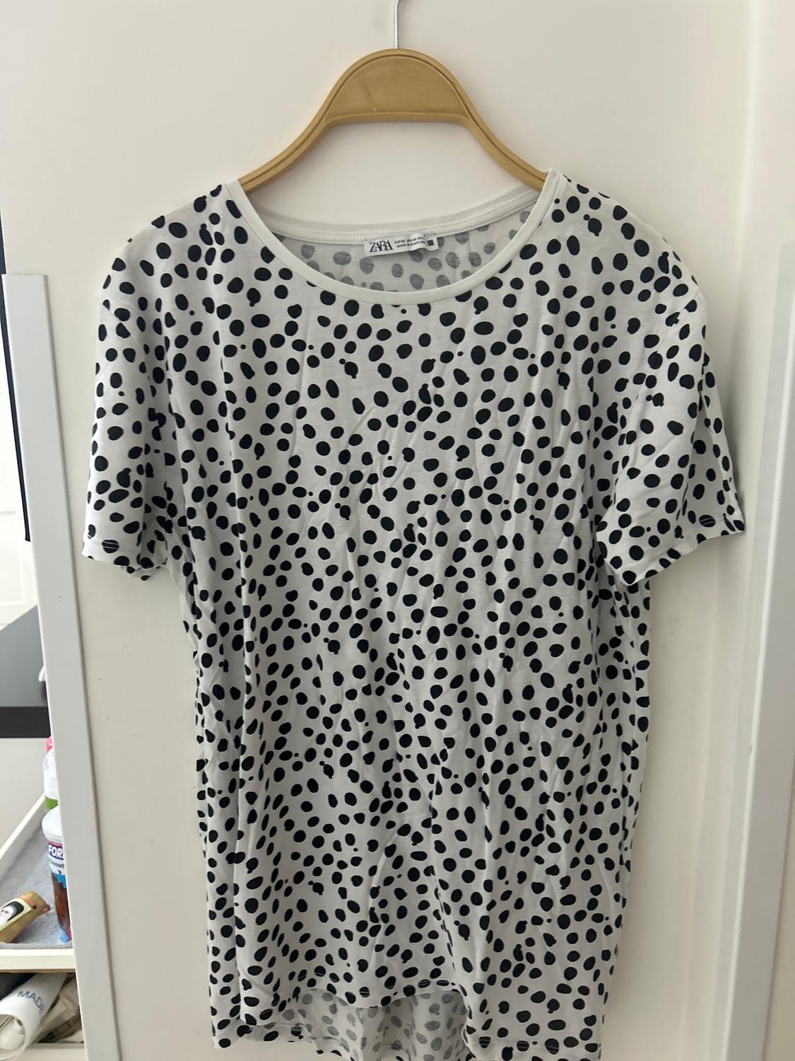 Λευκό t-shirt με στάμπα μαύρων πουά, κρεμασμένο σε ξύλινη κρεμάστρα πάνω από μια λευκή πόρτα.