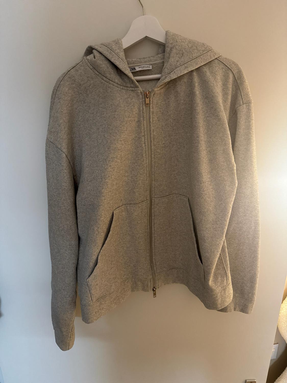 Grey zipped hoodie hanging on a white hanger against a light background. ZARA label visible on the neck.