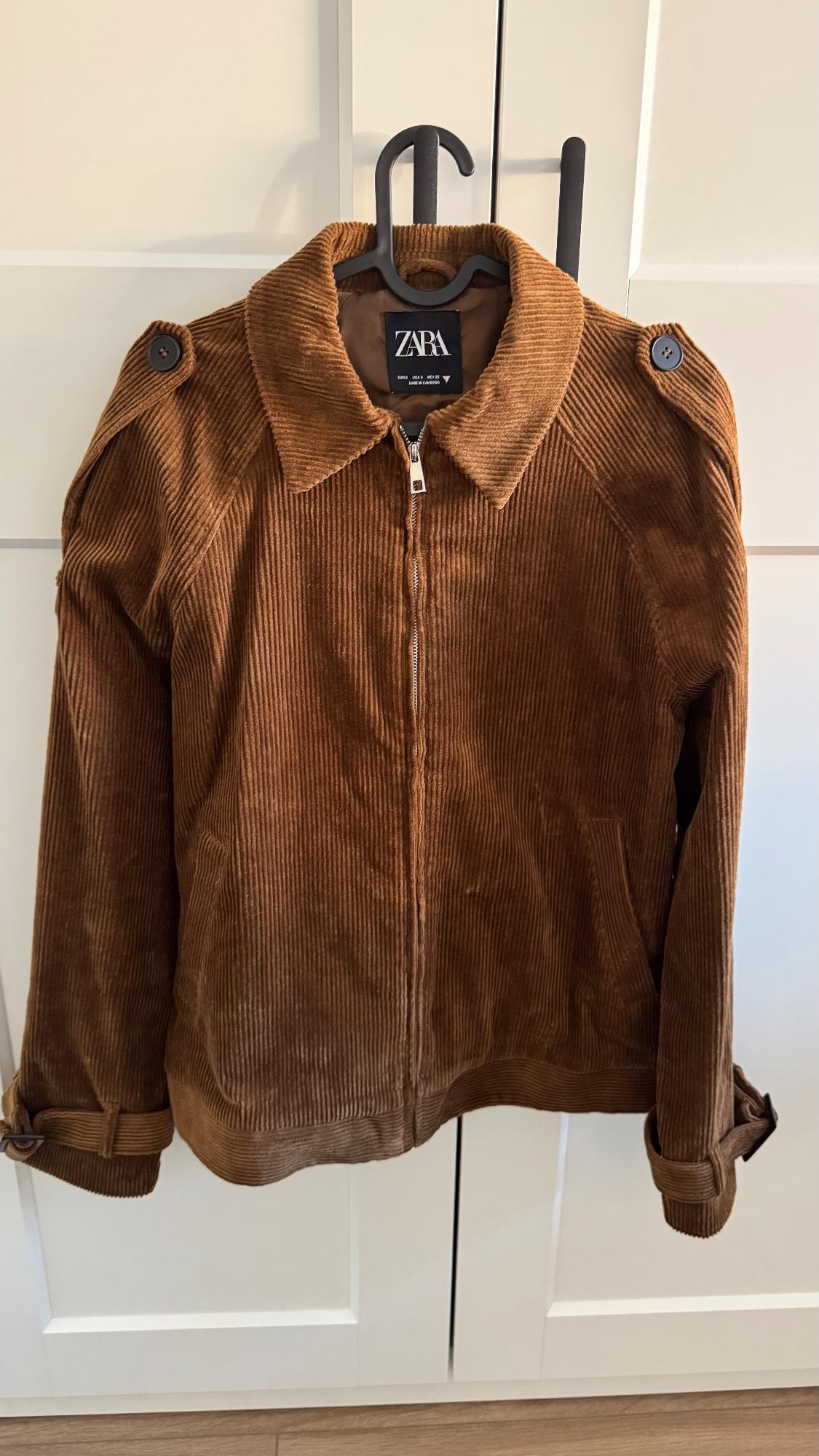 Brown corduroy jacket with zip and buttons on the shoulders, hanging on a black coat hanger. Visible label: ZARA. White background.