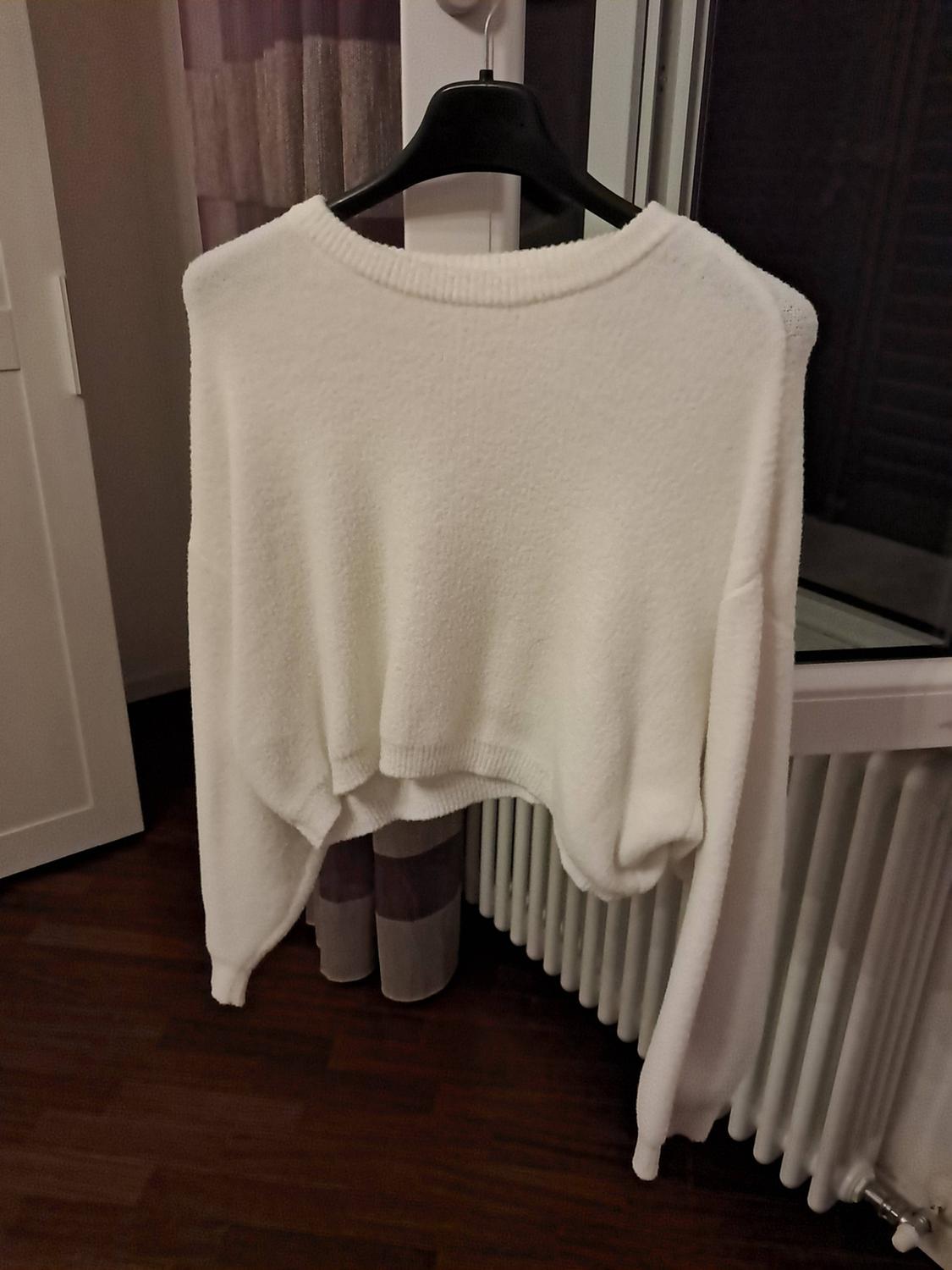 Short white jumper with a soft texture hanging on a black hanger in front of a window, above a white radiator and wooden floor.
