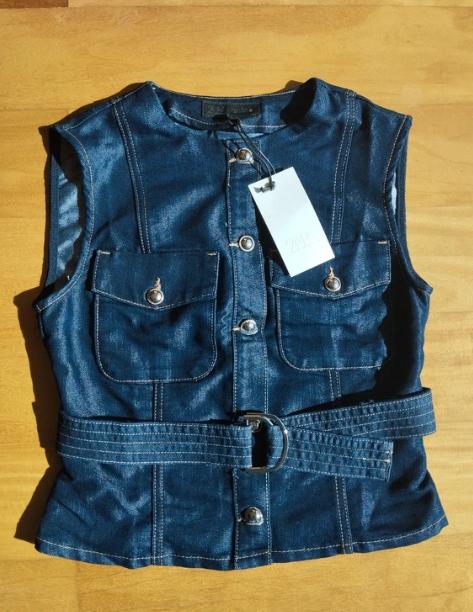 Sleeveless blue denim waistcoat with front pockets and belt, on a light wooden surface. White hanging tag.