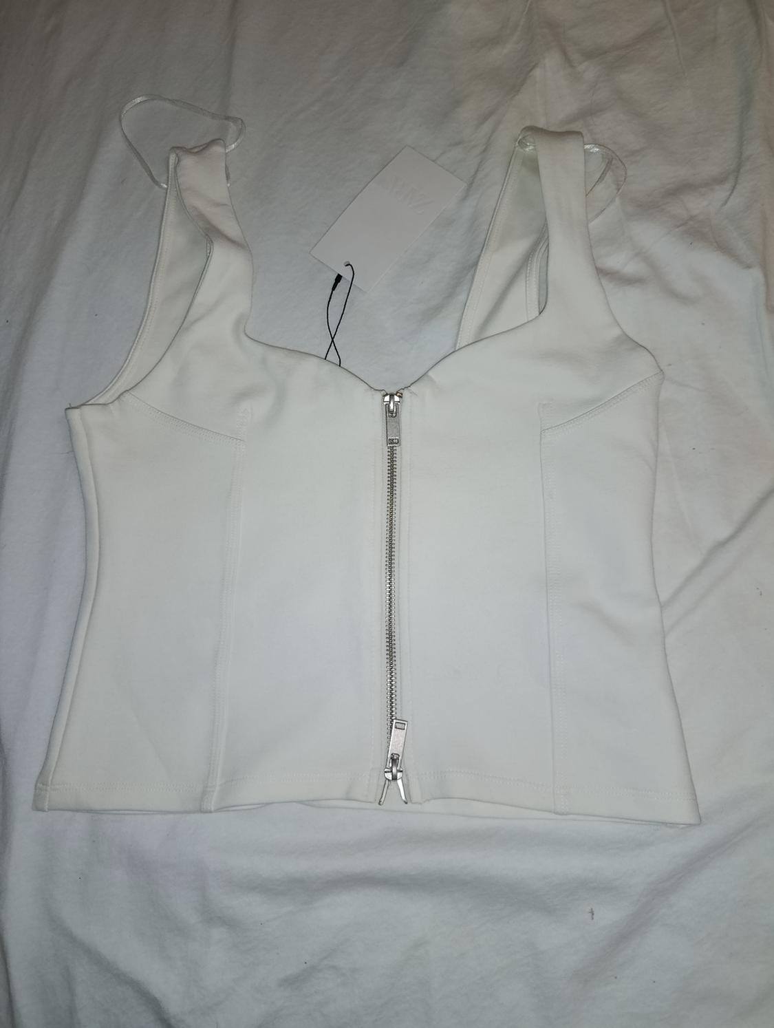 White strappy top with a silver front zip and visible stitching, laid on crumpled white fabric.