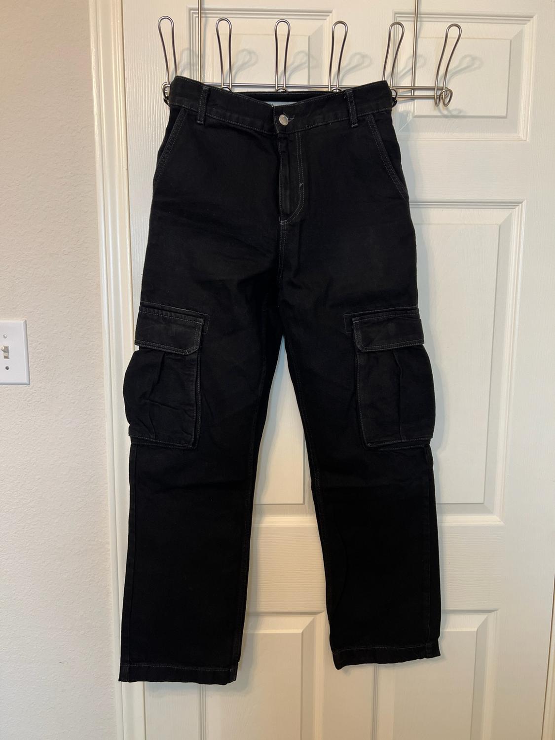 Black cargo trousers with side pockets, hanging on a metal hanger over a white door indoors.