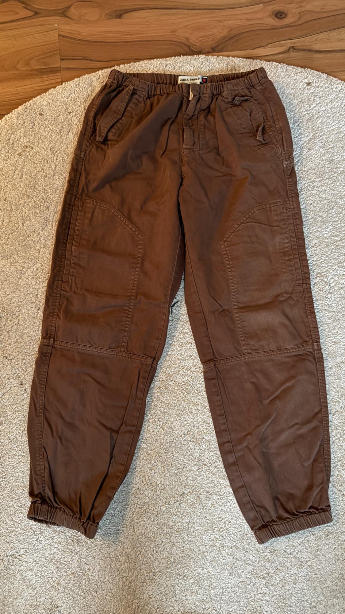 Brown trousers with an elasticated waist and pockets, on a beige rug and wooden floor. Visible label: "ZARA SRPLS".