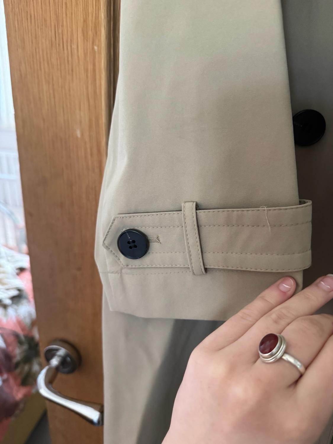 Here is the rephrased sentence, maintaining the same meaning and context: "Close-up of the sleeve of a beige trench coat featuring a strap and a black button. A hand wearing a red ring brushes against the cuff. In the background, a wooden door is visible."