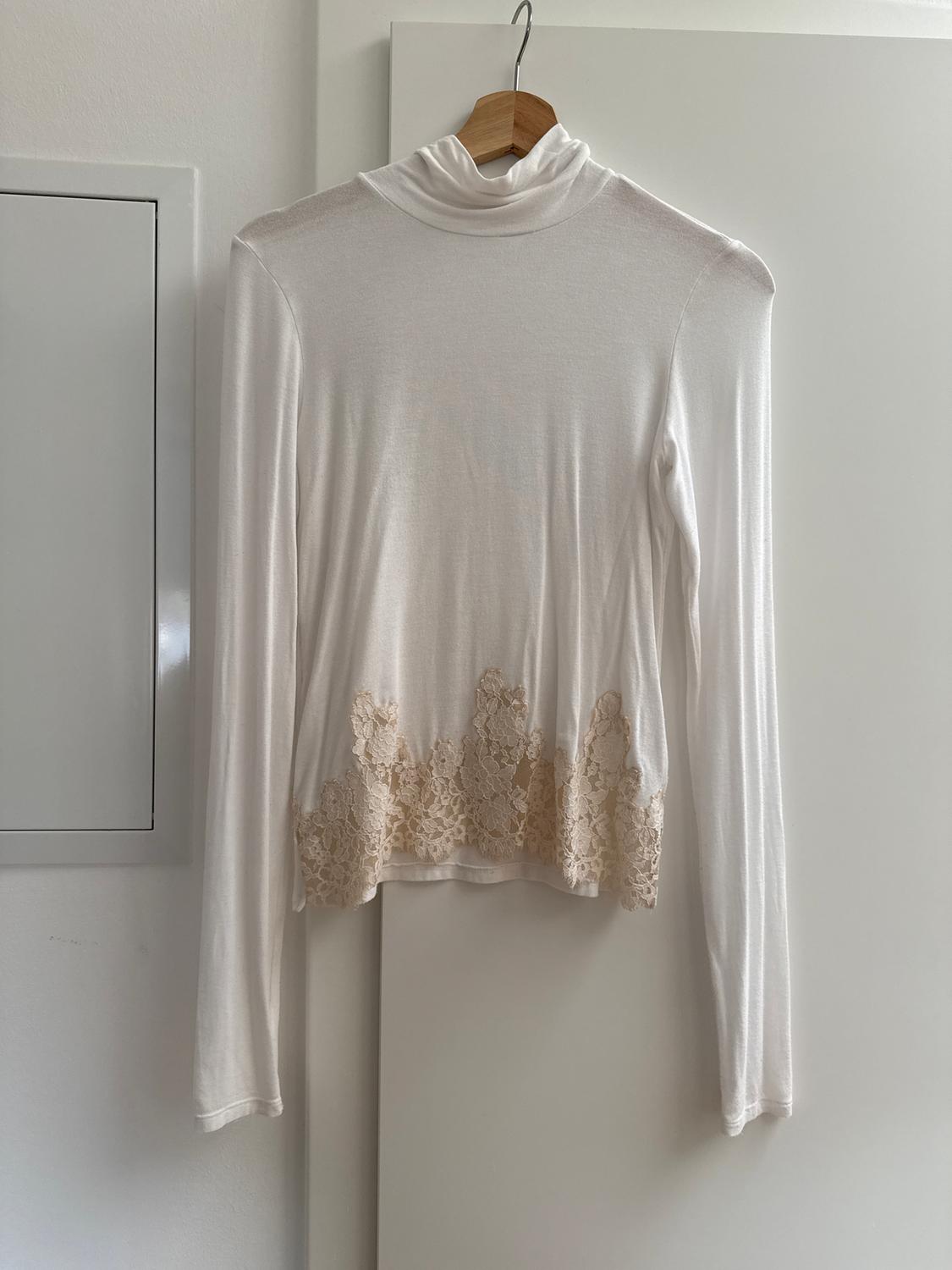Long-sleeved white blouse with a high neck and beige lace details on the hem, hanging on a wooden hanger.
