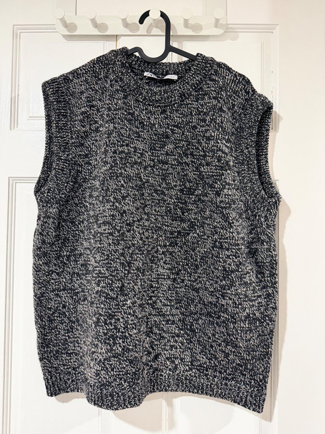 Grey and black knitted waistcoat hanging on a black hanger over a white door. Casual and cosy style.