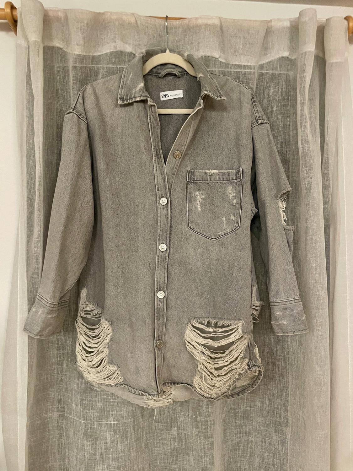 Distressed gray denim shirt with rips at the hem and pocket, hanging on a hanger in front of a white curtain.