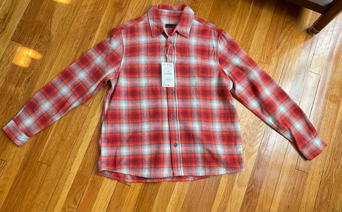Long-sleeve shirt with a plaid pattern in red, white, and blue tones, laid out on a wooden floor, with a price tag.