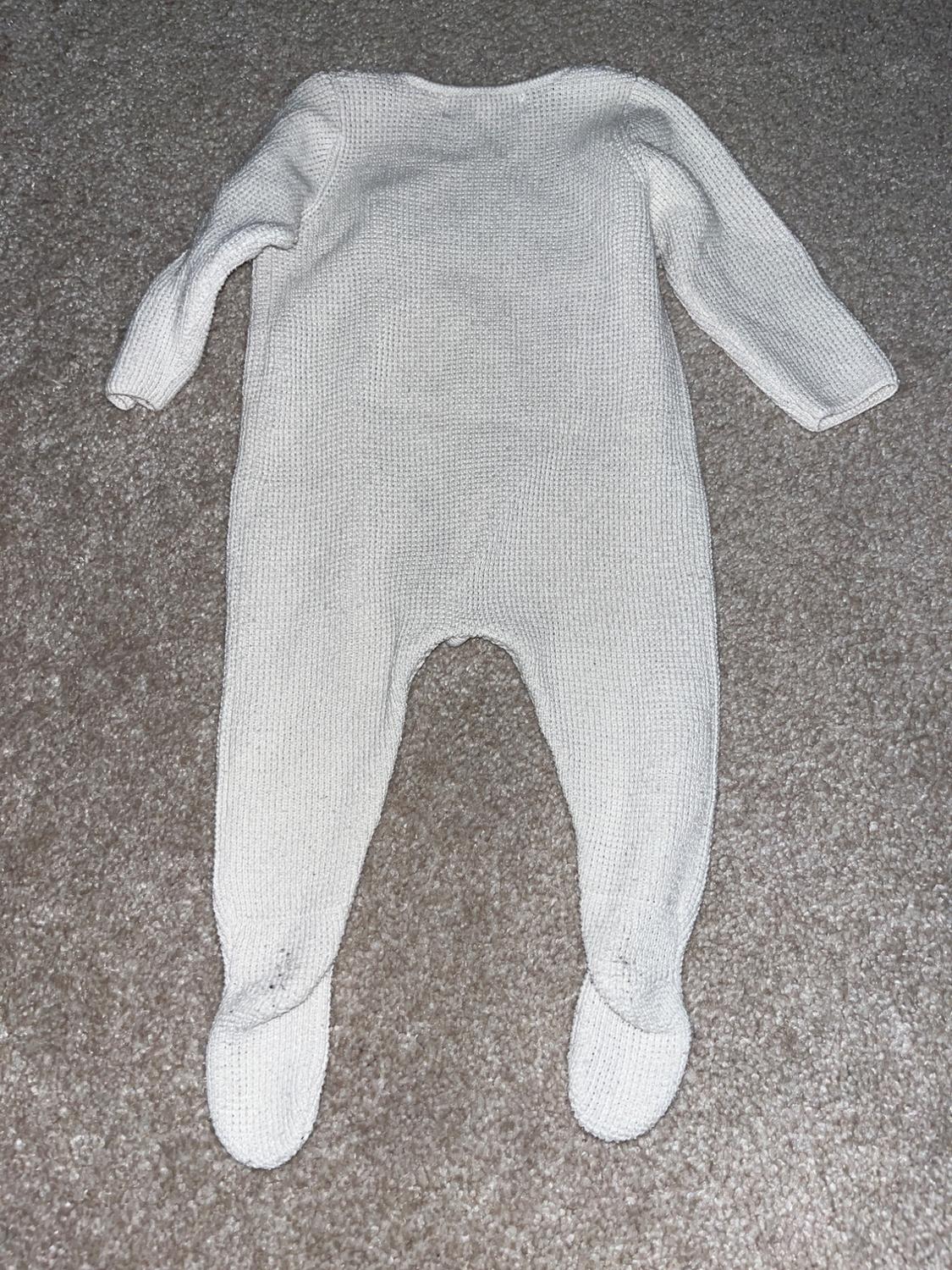 White knitted romper with long sleeves and covered feet, laid out on a grey rug.