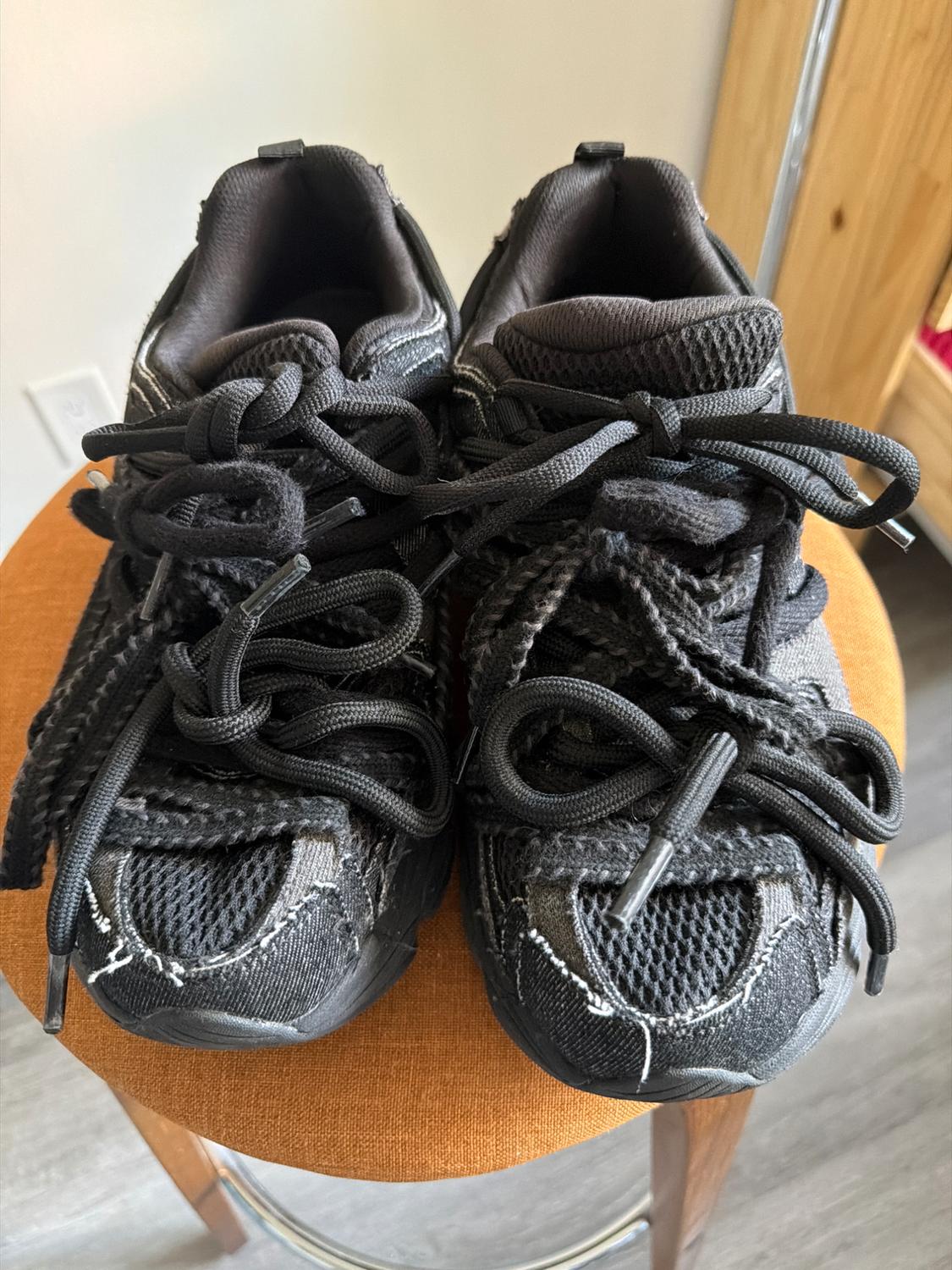 Black trainers with thick laces and visible stitching, placed on an orange stool in an indoor setting.