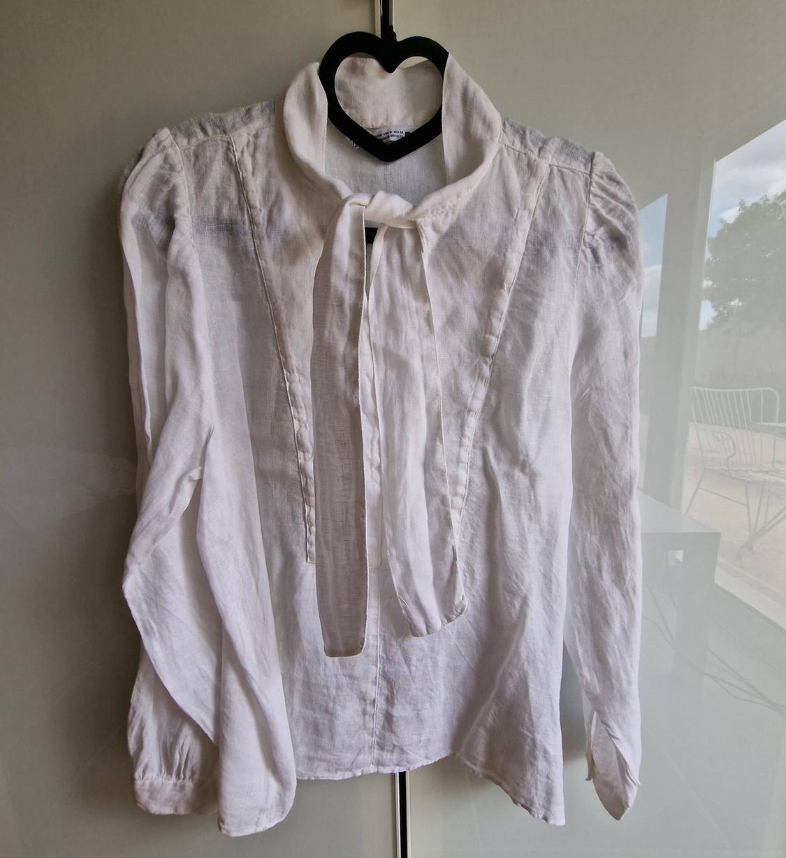 White long-sleeved blouse with a neck tie, hanging from a black heart-shaped hanger in front of a reflective glass.