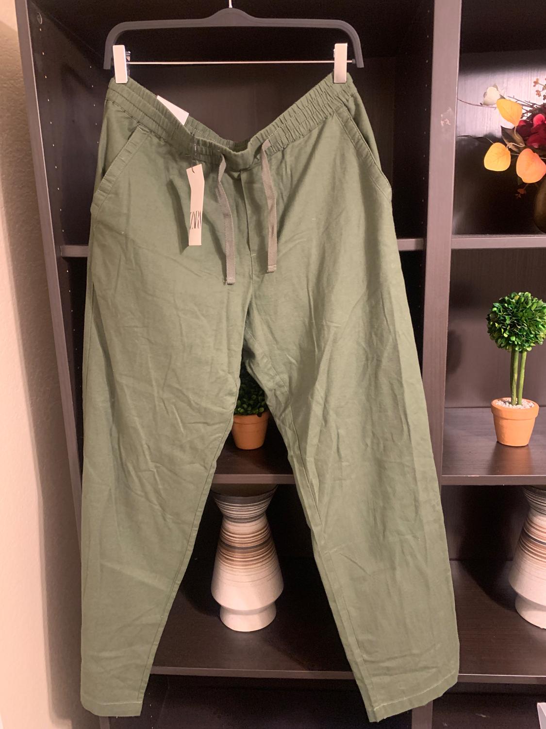 Green trousers hanging on a hanger inside a dark shelf, with a visible label. Decorated with plants and vases around.