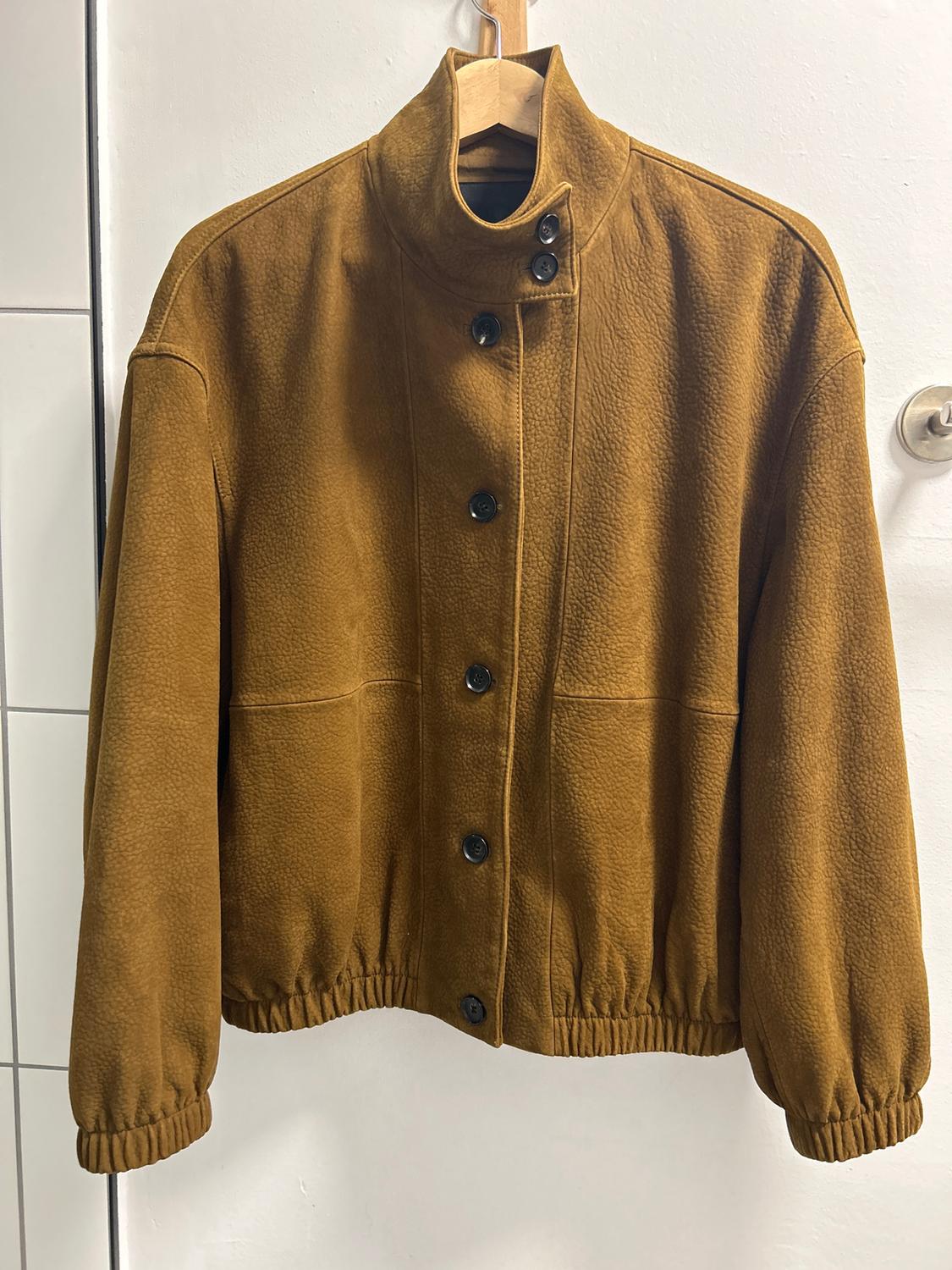 Brown jacket hanging on a wooden hanger, with a high collar and black buttons. White background and tiles to the left.