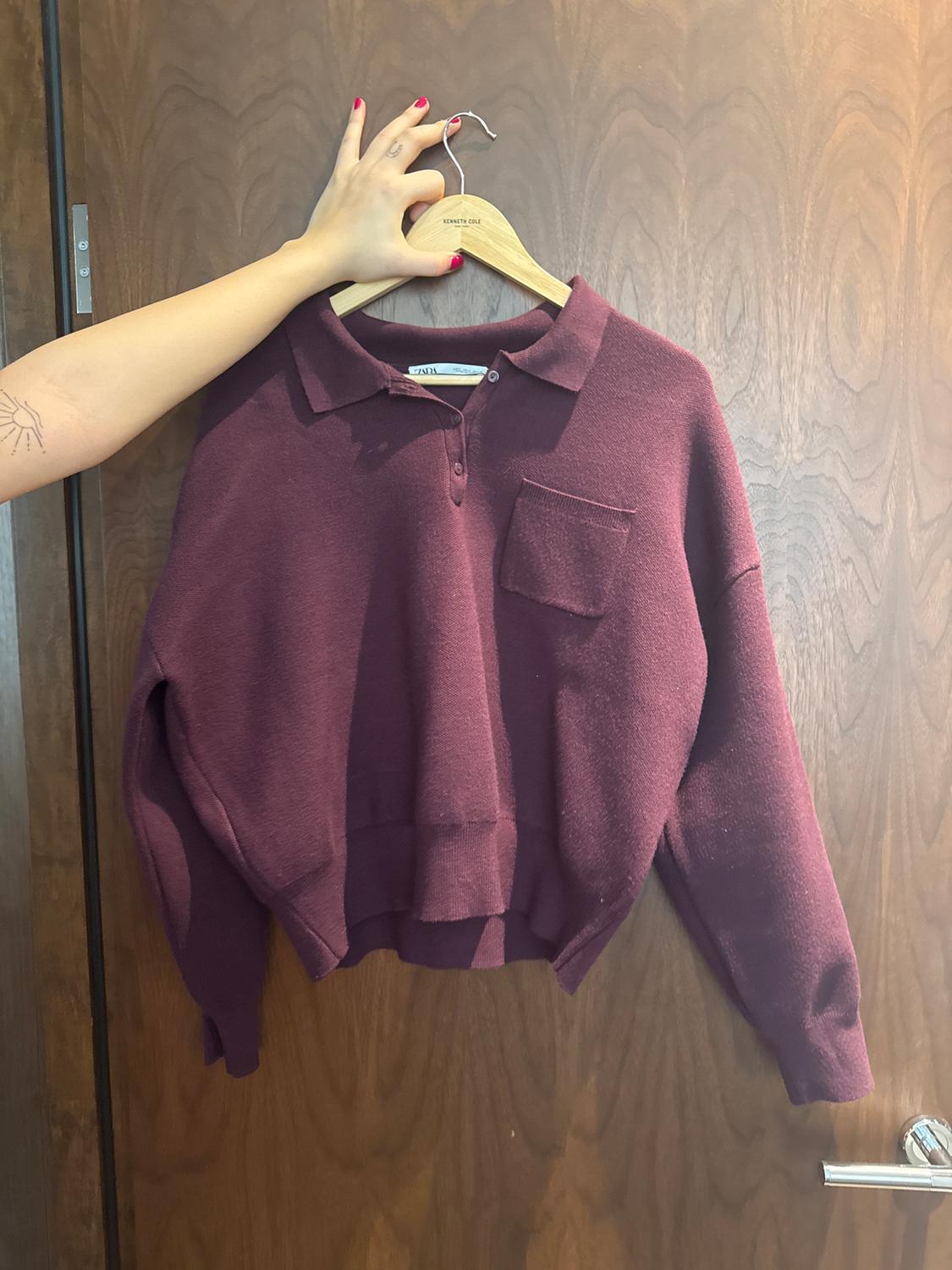 Burgundy polo neck jumper with a pocket, hanging on a wooden hanger. Hand with red nails holding the hanger. Door in the background.
