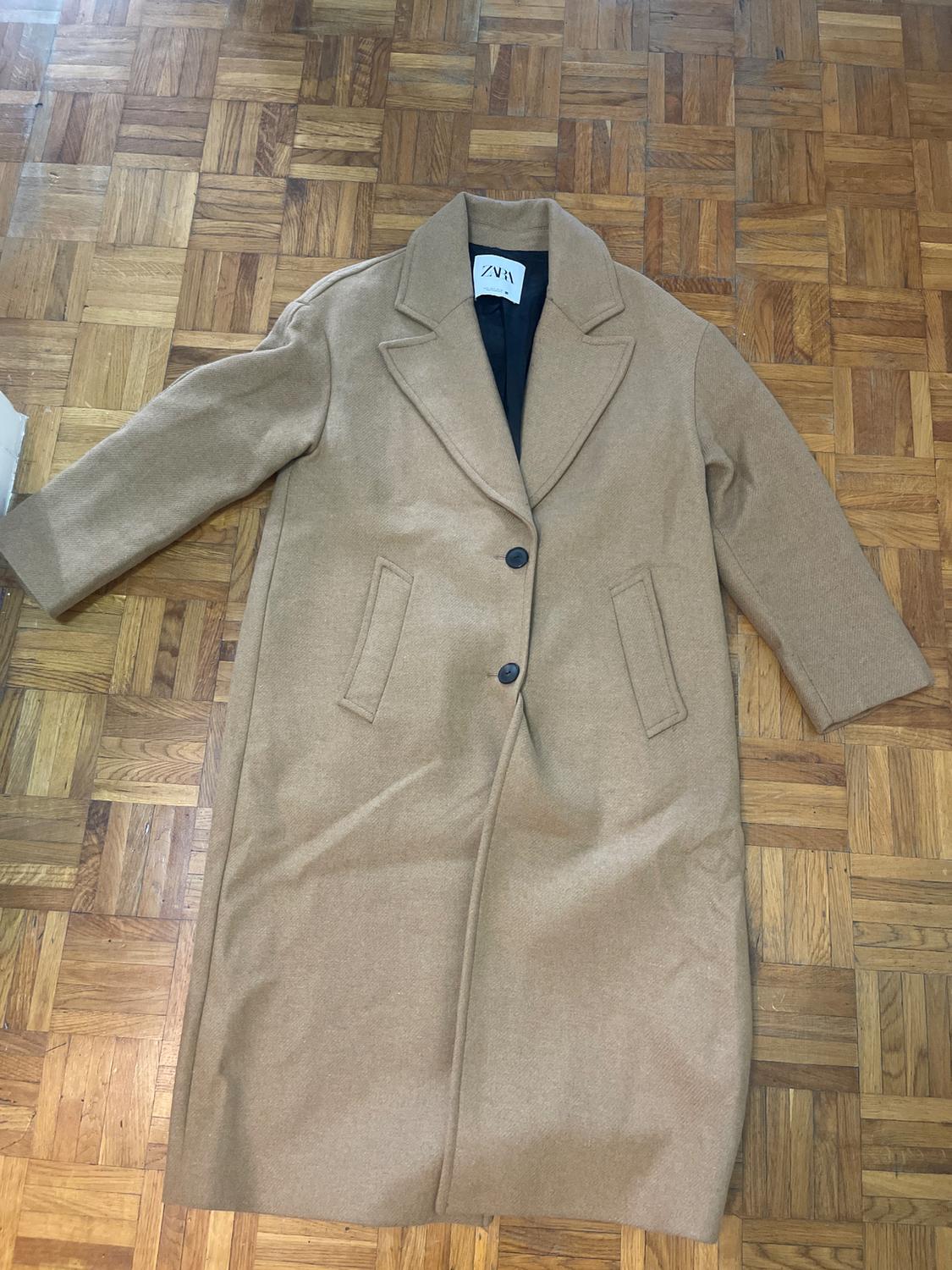Long camel coat with lapels and two buttons, on a wooden floor. Visible label with text "ZARA".