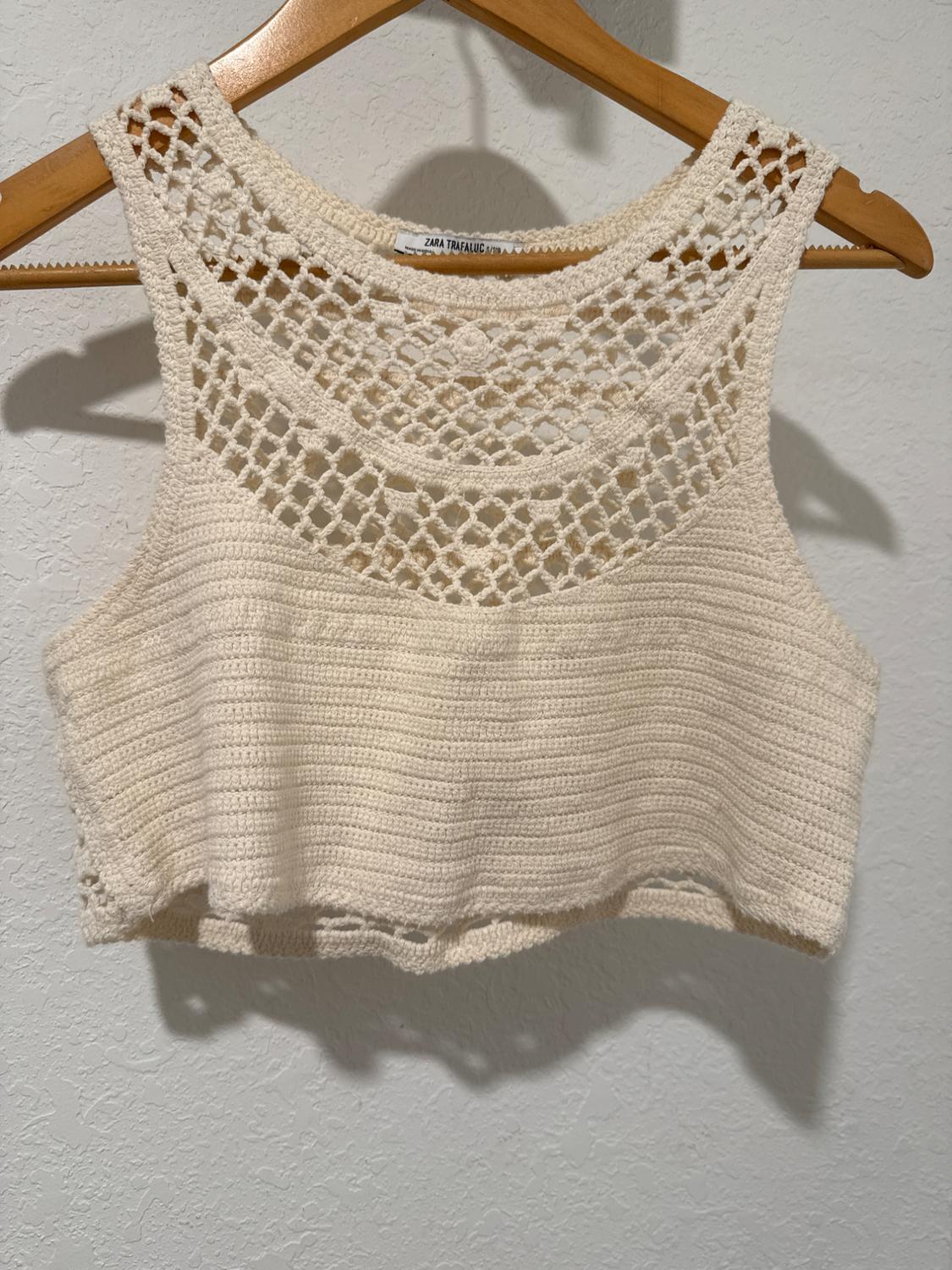 Beige crochet top hanging on a wooden hanger. The design features an open-knit pattern at the top.