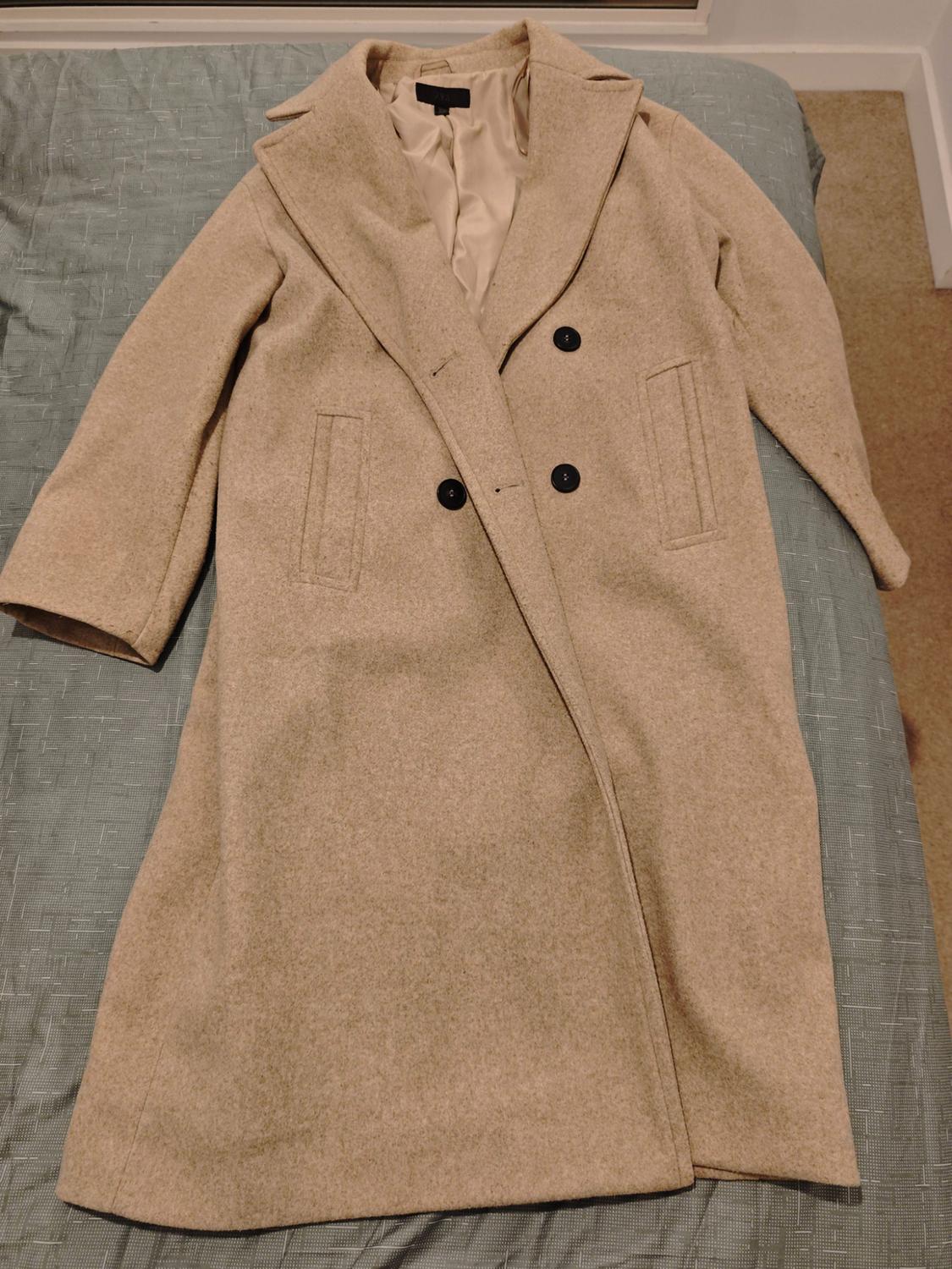 Beige long coat with dark buttons and wide lapels, draped over a bed with blue sheets.