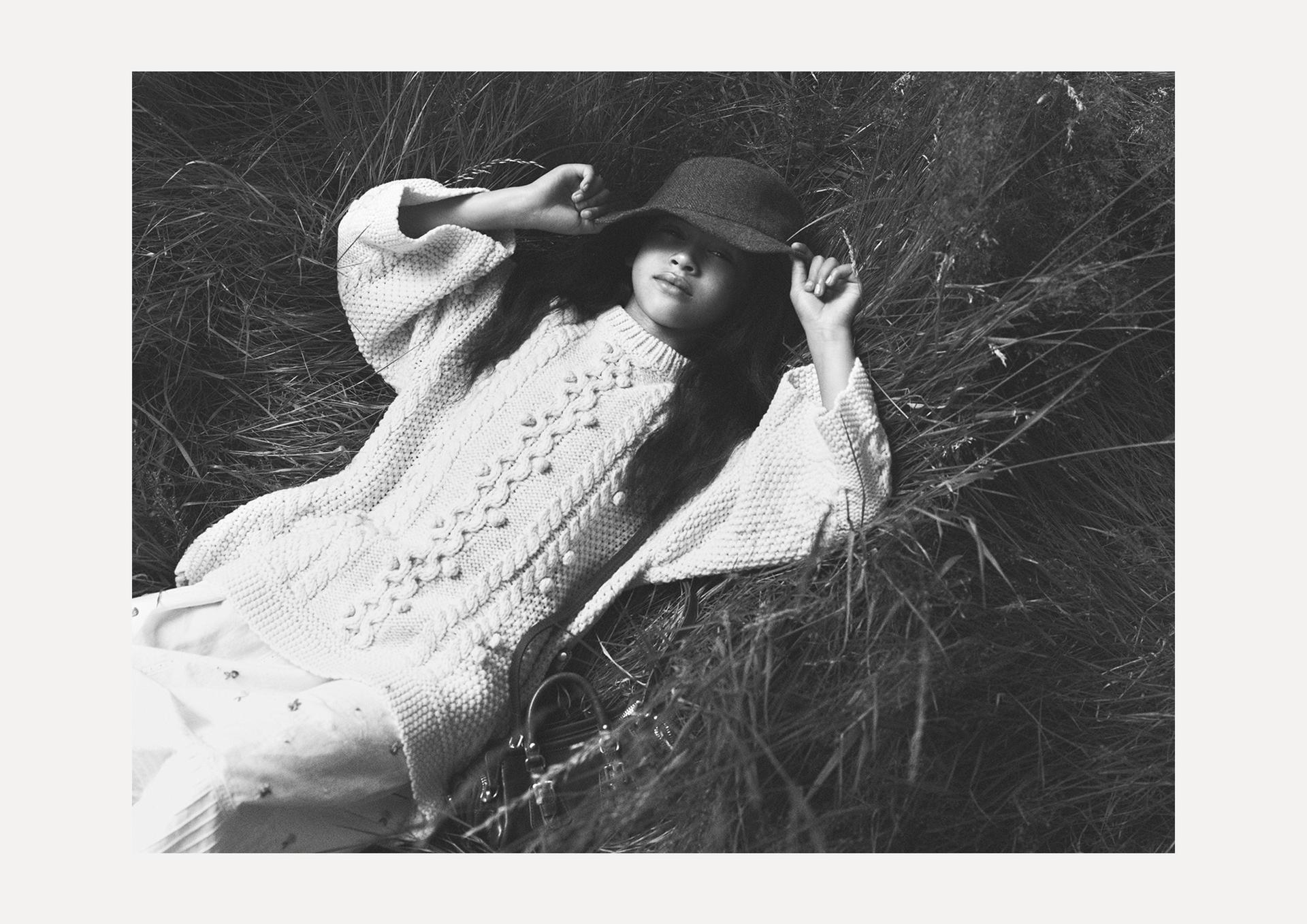 Black and white: model reclining on grass, wearing a chunky knit jumper and hat, creating a relaxed and natural atmosphere.