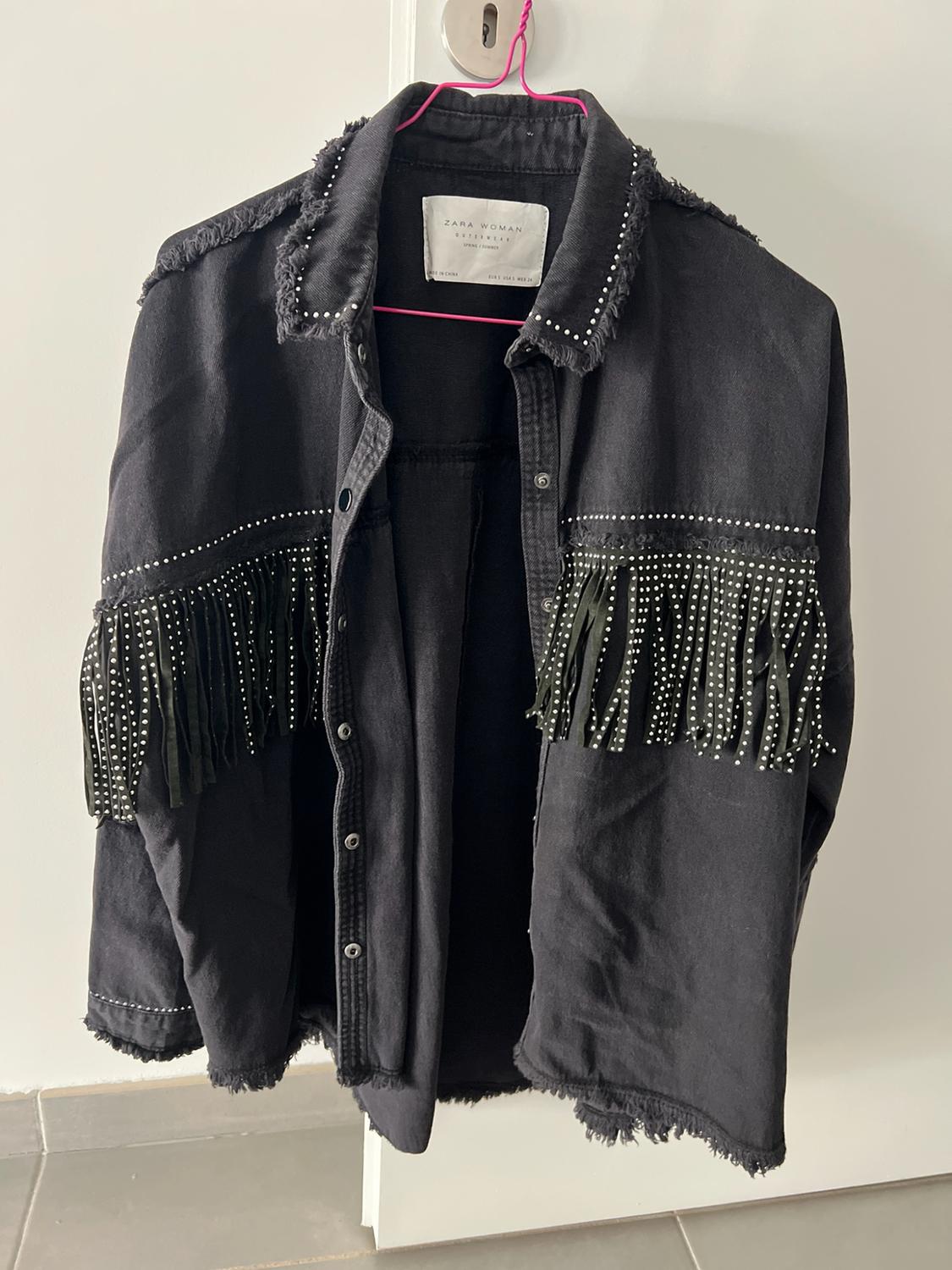 Black jacket with fringing and studs, hanging on a pink hanger on a white door. Visible label: ZARA WOMAN.