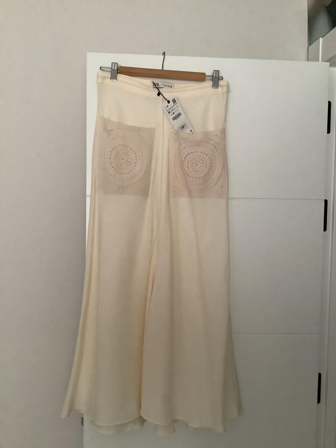 Cream wide-leg trousers with circular crochet pockets, hanging on a wooden hanger on a white door. Label with the text ZARA.