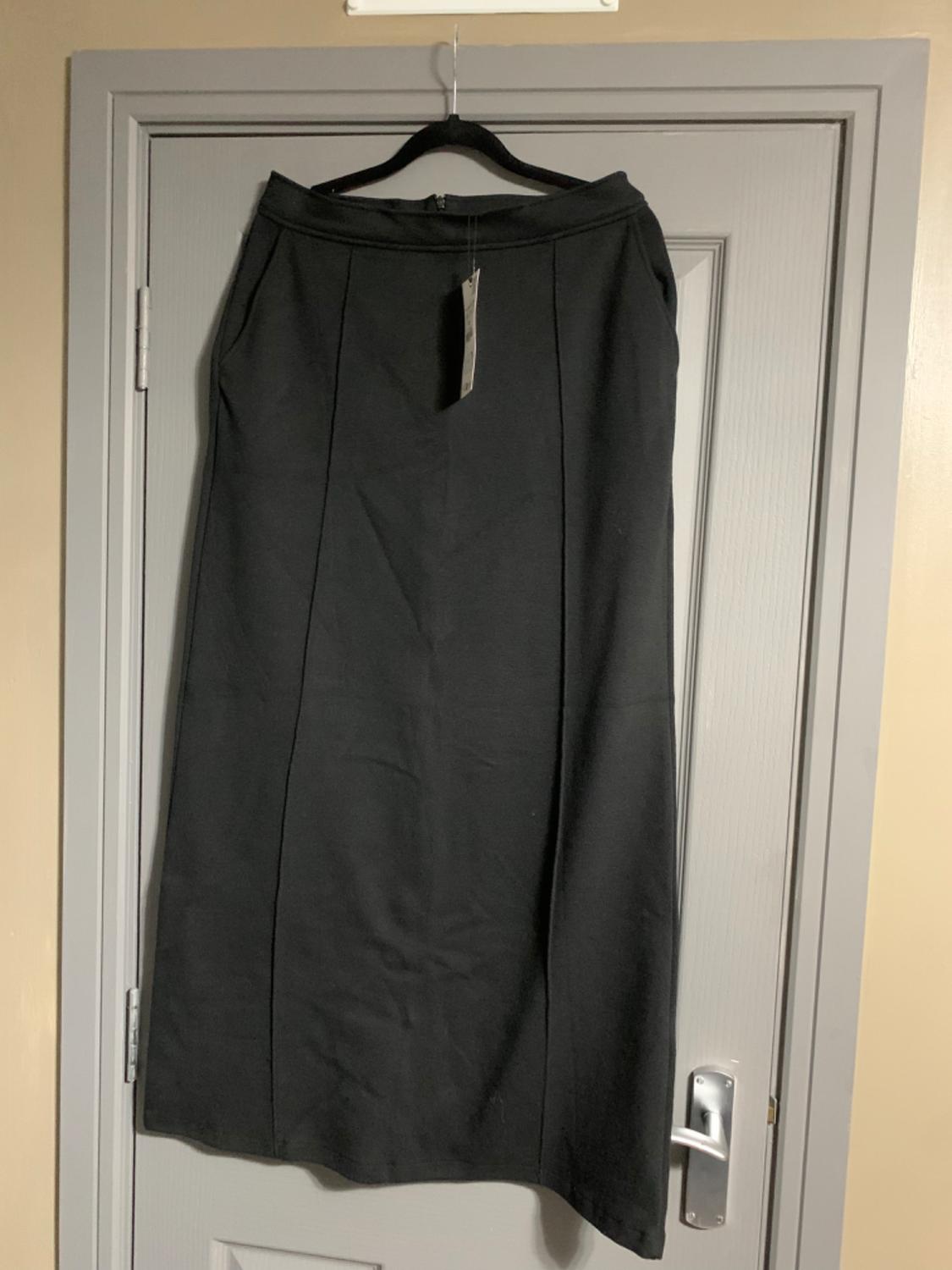 Long black skirt hanging on a hanger over a grey door. The skirt has a simple design with vertical seams.