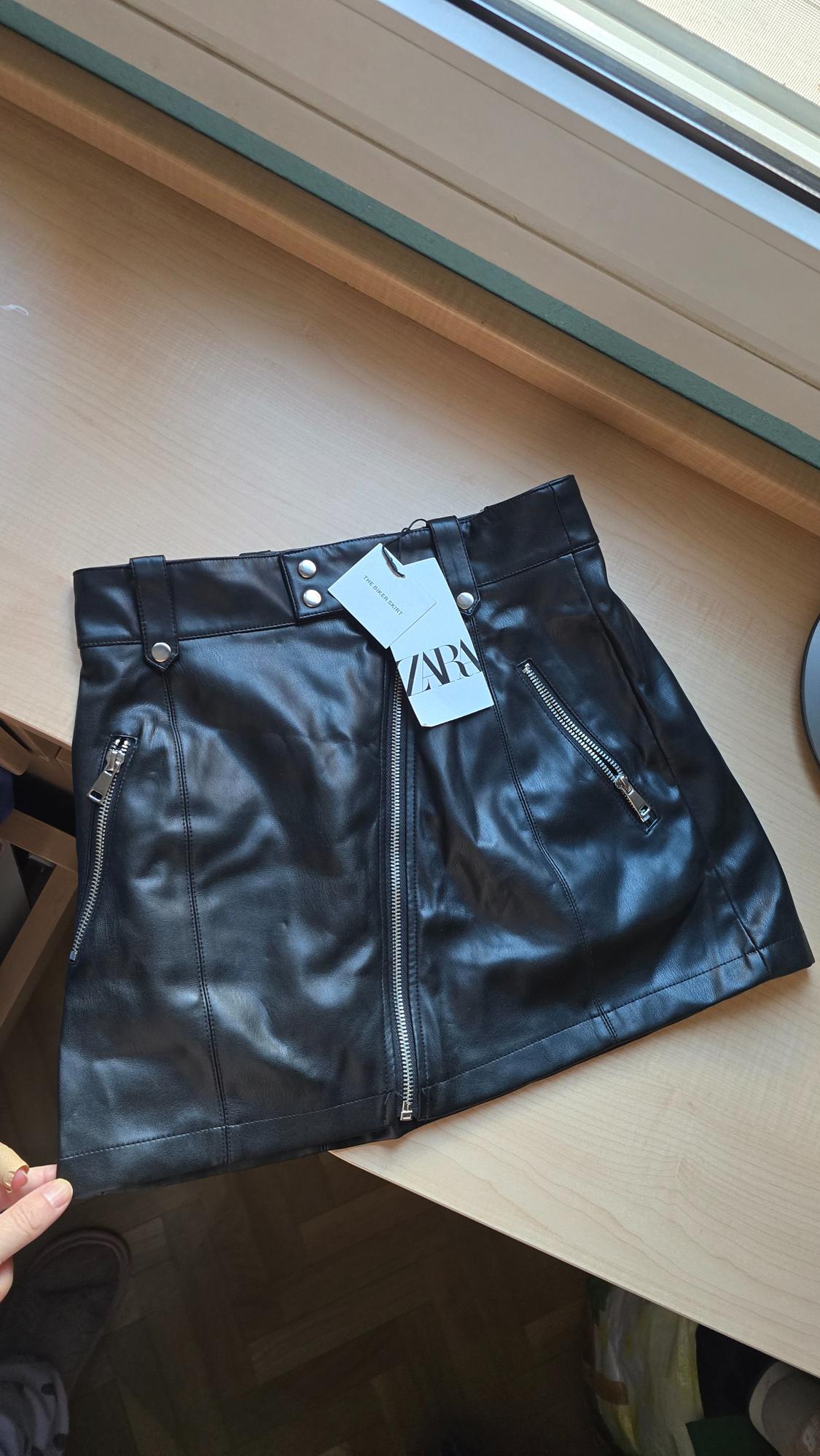 Black synthetic skirt with zips and Zara tag on a light wooden table next to a window.