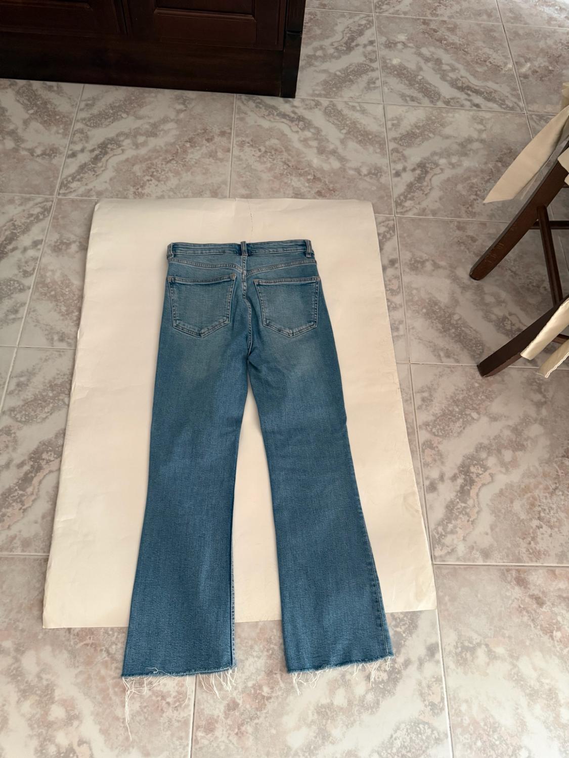 Blue jeans with frayed hems on white paper on a tiled floor. Relaxed composition with wooden furniture in the background.