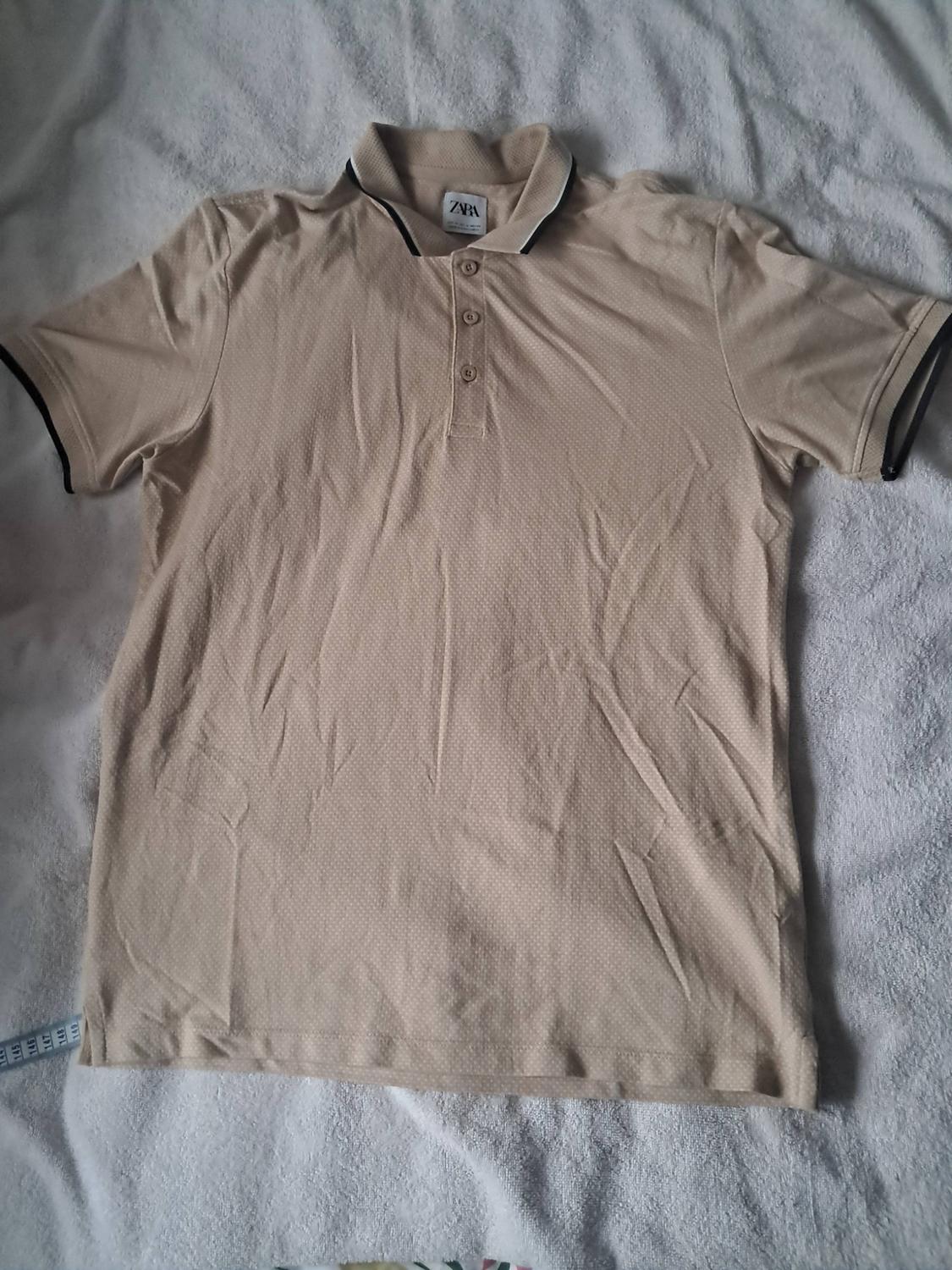 Beige short-sleeved polo shirt with dark trim on the collar and sleeves, laid flat on a white textured surface. ZARA label visible.