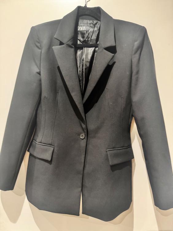 Black women's jacket hanging on a hanger, with classic lapels and one front button. Visible label on the inside.