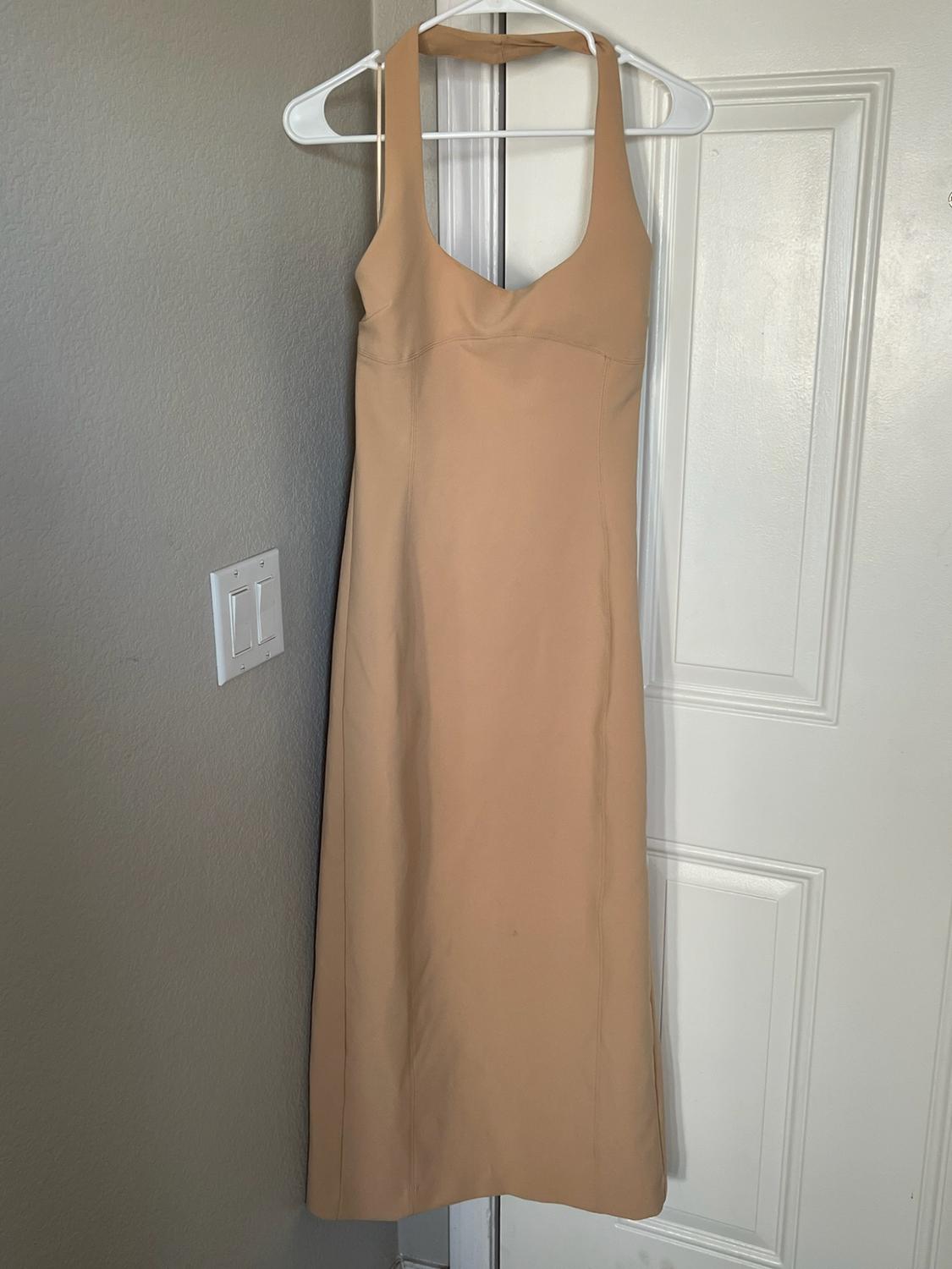 Beige maxi dress with a halter neck hanging on a white hanger on a white door, next to a gray wall with a light switch.