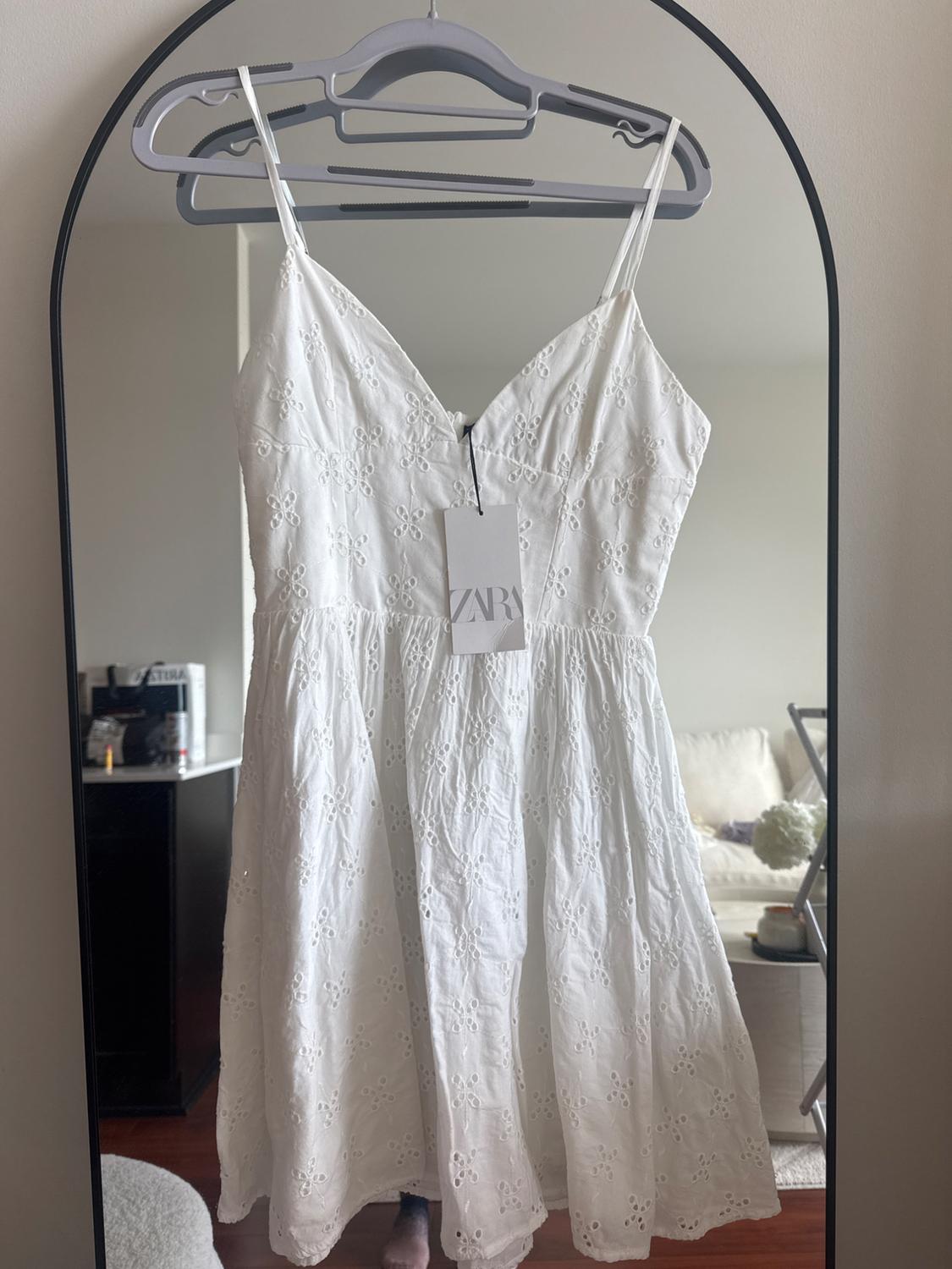 White dress with thin straps and floral embroidery hanging on a hanger in front of a mirror. Visible label with text "ZARA".