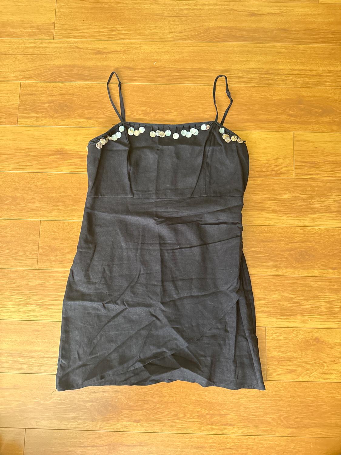 Black spaghetti strap dress with circular embellishments on the neckline, on a light wooden floor.