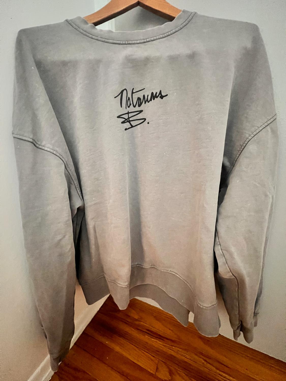 Gray sweatshirt hanging on a wooden hanger, with a black signature on the back. White wall background and wooden floor.