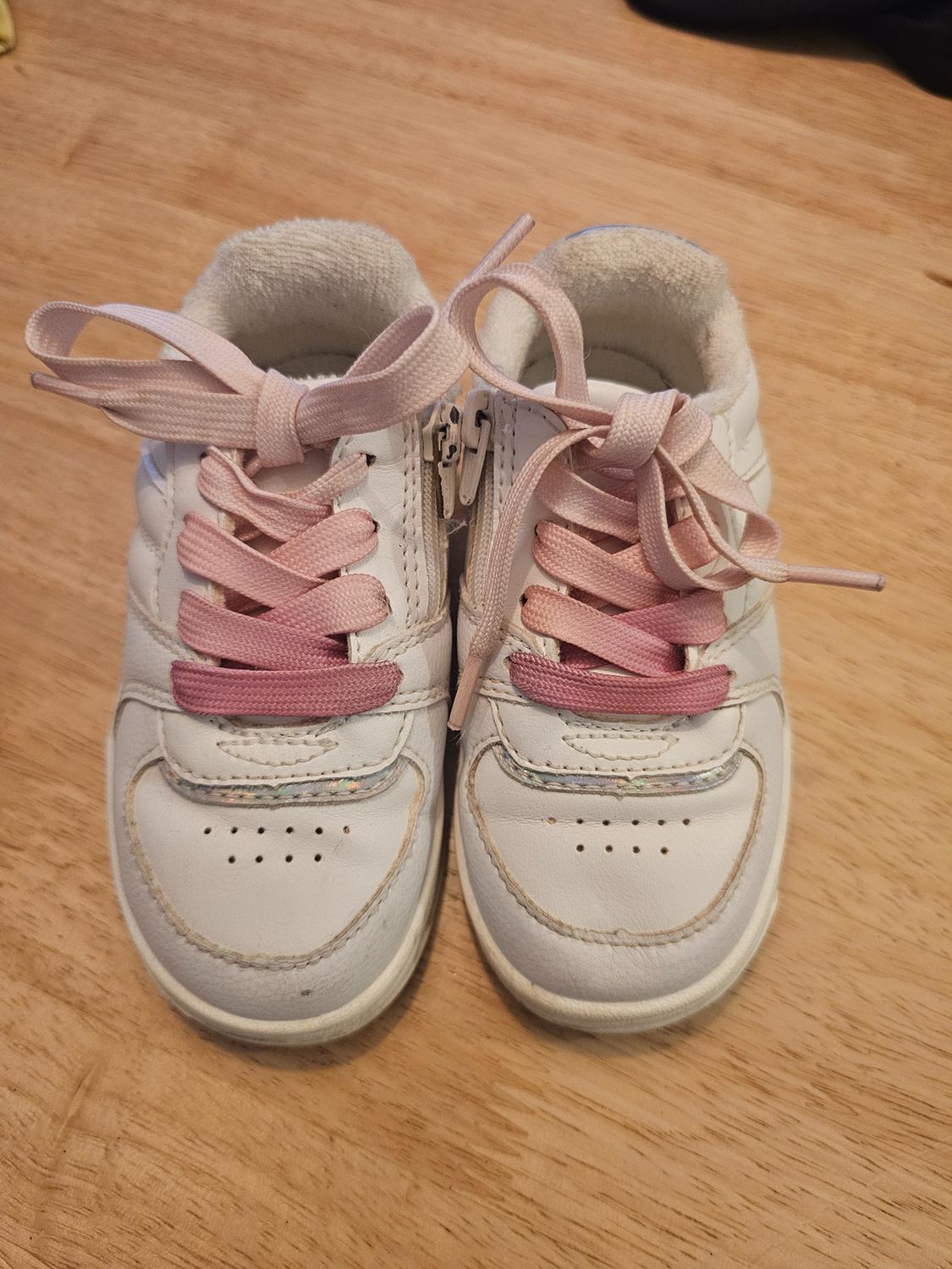 White children's trainers with pink laces on a light wooden surface.