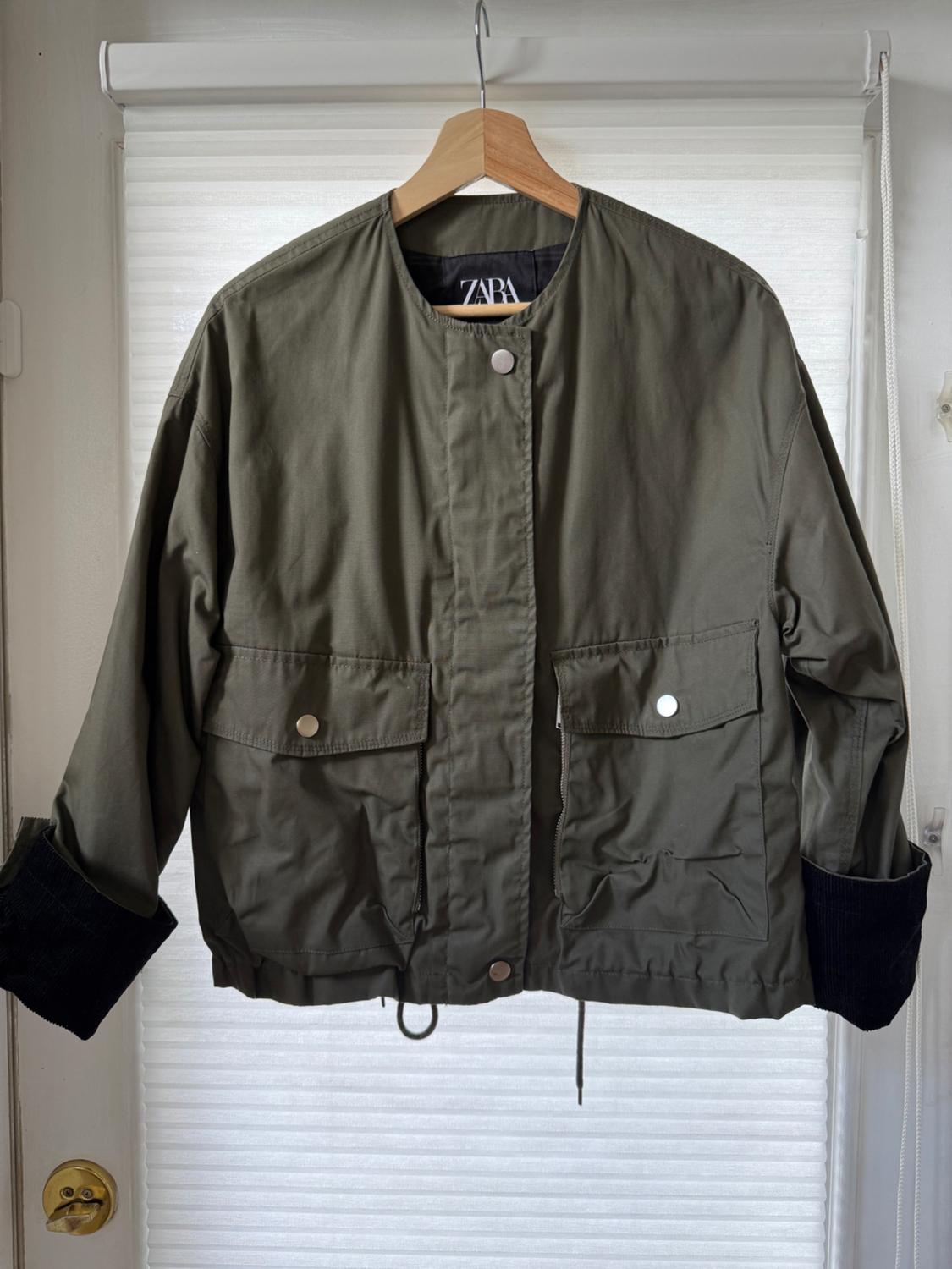 Olive green, long-sleeved jacket with front pockets and dark cuffs, hanging on a wooden hanger. Visible tag: ZARA.