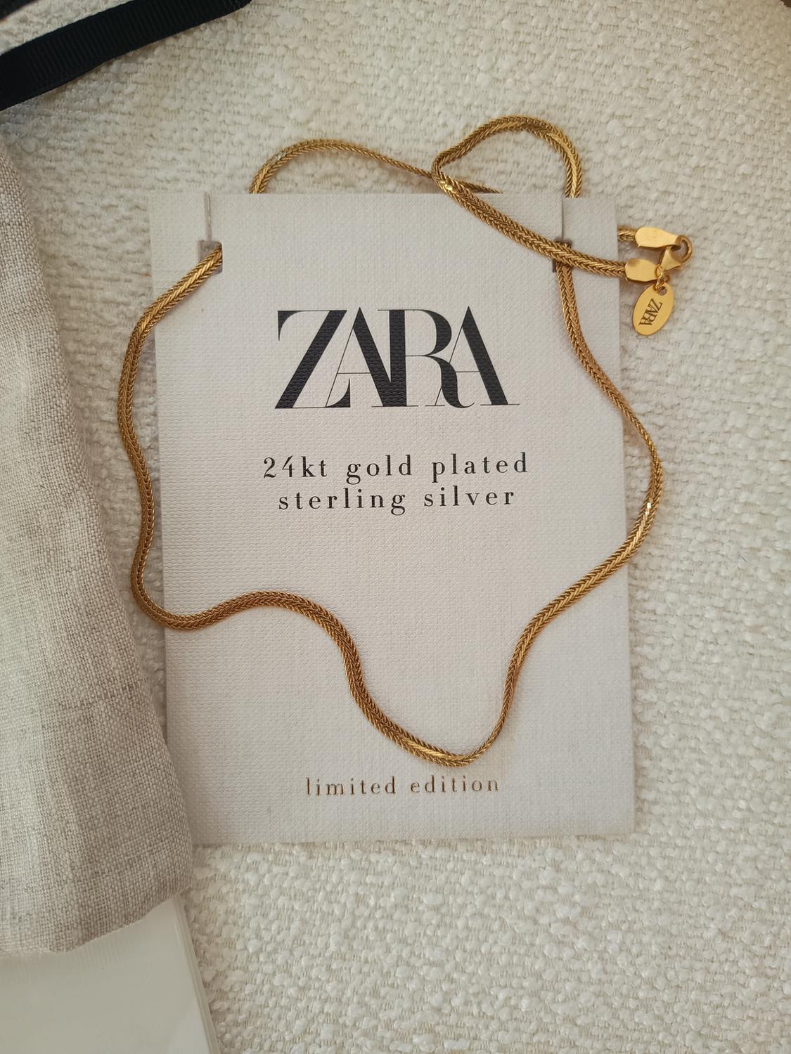 Gold chain on card with text: "ZARA, 24kt gold plated sterling silver, limited edition". Beige fabric background.