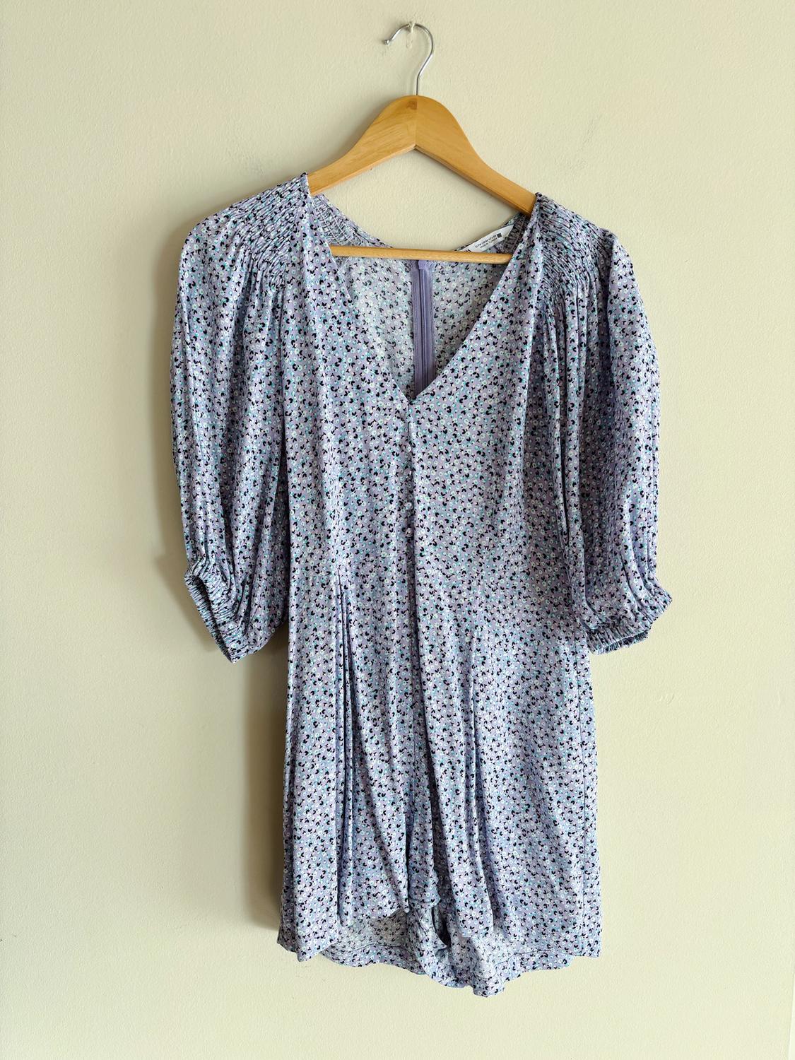 Lightweight fabric playsuit with a floral print in blue and lilac tones, hanging on a wooden hanger against a beige wall.