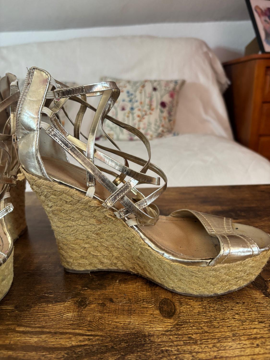 High wedge sandal with metallic gold straps and esparto sole on a wooden table, bed background with floral cushion.