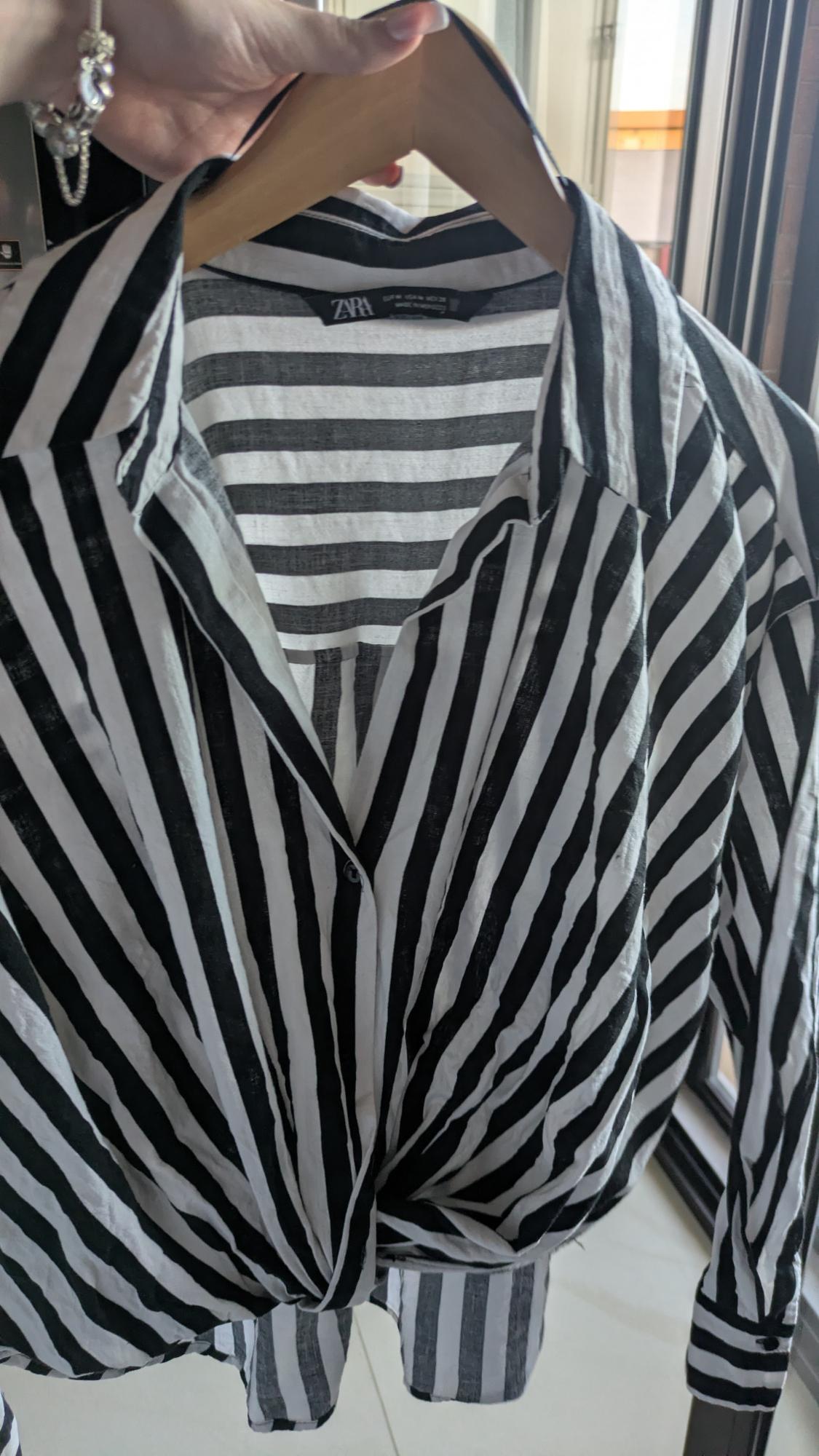 Hand with bracelet holding a hanger with a black and white striped shirt knotted at the hem. ZARA label. Bright window background.