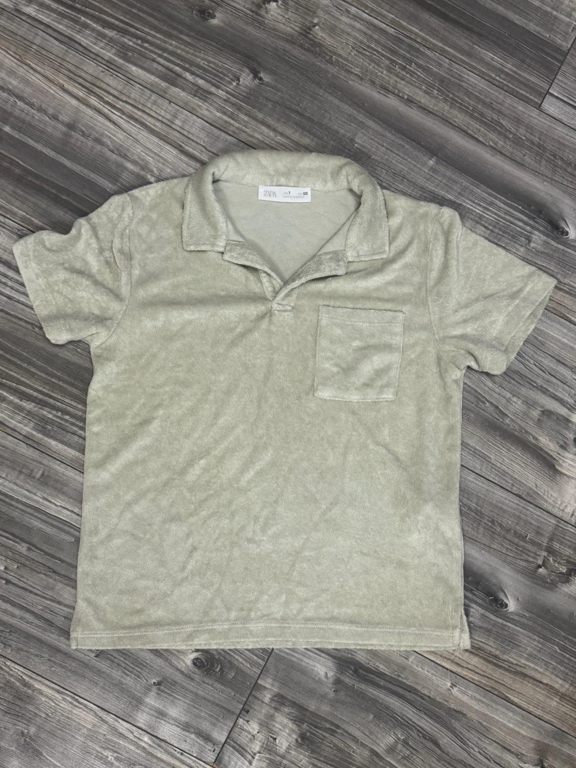Light green short-sleeved textured polo shirt with a collar and chest pocket. Label with ZARA text. Garment against a grey wooden background.