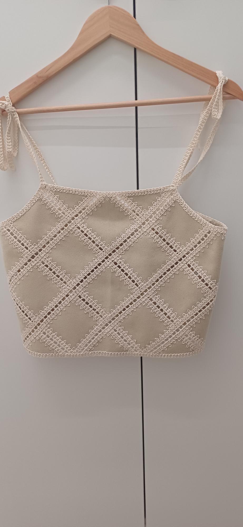 Beige crop top hanging on a wooden hanger, with a white lace diamond pattern and thin tied straps. White background.