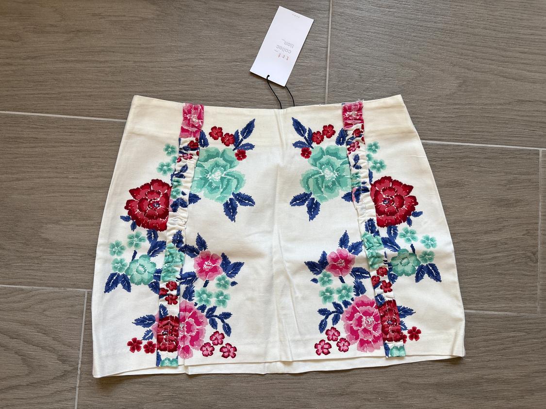 White mini skirt with floral print in shades of red, green and blue, vertical ruched detailing and hanging tag on a grey floor.
