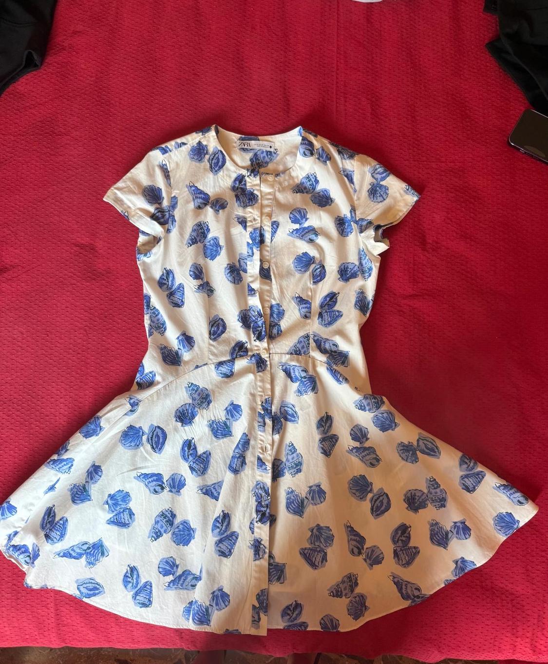 White short dress with blue shell print and front buttons, laid out on red fabric. Label with text ZARA.