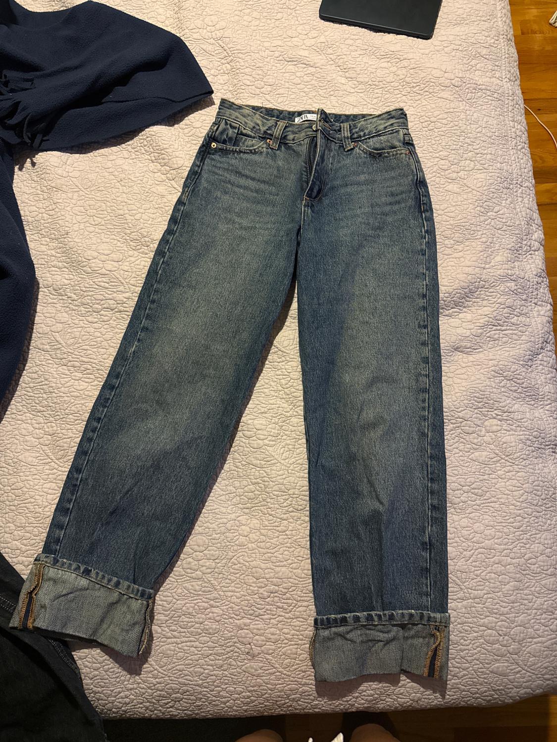 Straight-leg blue jeans with a hem, placed on a white bedspread. To the left, a dark garment.