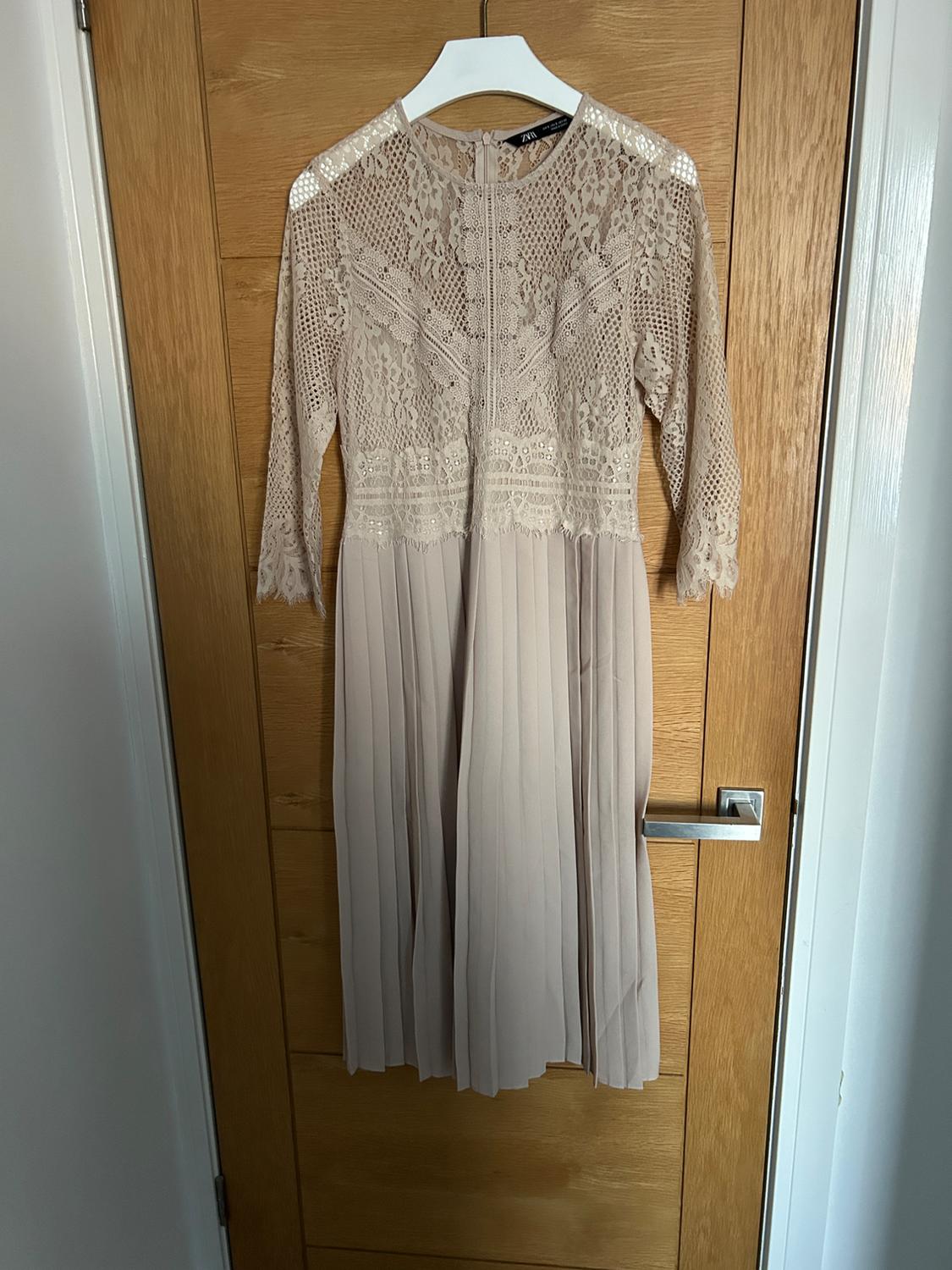 Beige dress hanging on a hanger, with lace top and three-quarter sleeves, pleated skirt, light wooden door background.