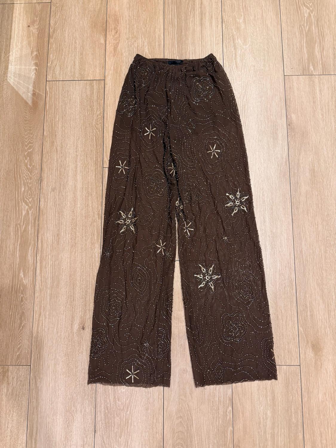 Semi-sheer brown wide-leg trousers with sparkly flower and star embroidery, laid out on a light wood floor.