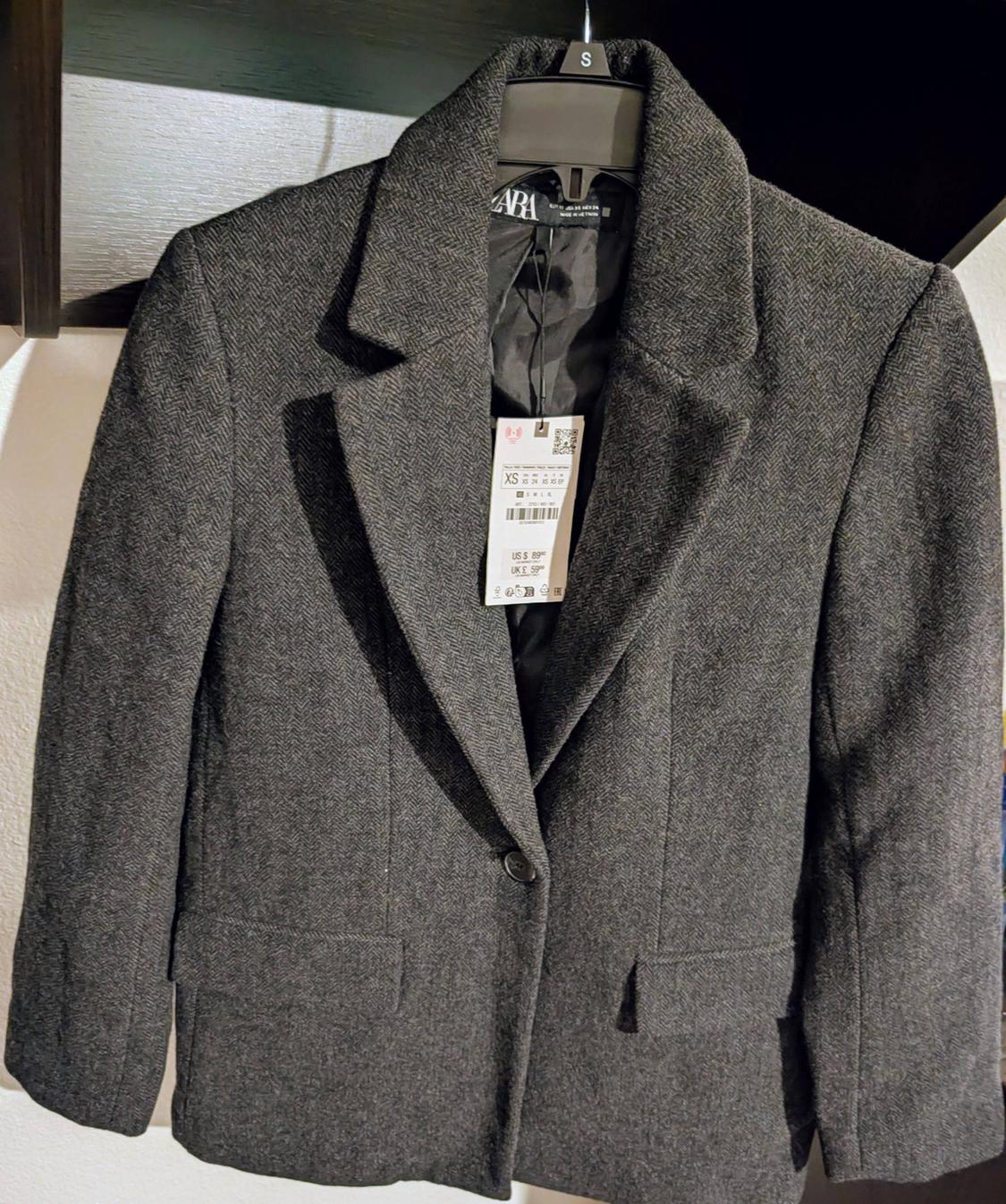 Gray wool jacket hanging on a black hanger with a visible tag. White wall background and dark shelf.