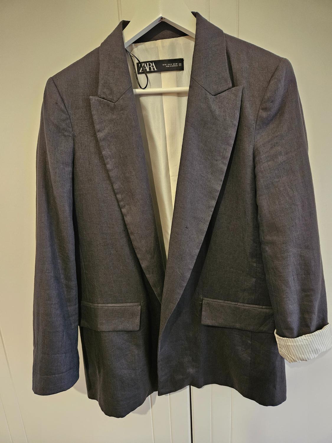 Grey ZARA jacket hanging on a white hanger, with wide lapels and front pockets, against a light background.