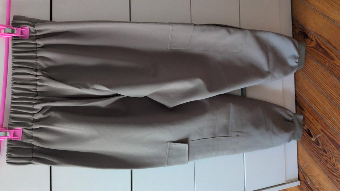 Beige cargo trousers with an elasticated waist and pockets, hanging on a pink hanger against a background of white panels and a wooden floor.